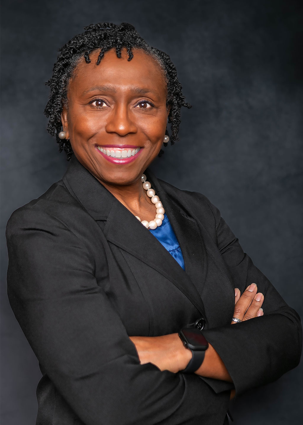 Portrait of a smiling woman with short curly hair, wearing a black blazer, pearl necklace, and a blue top, crossing arms.