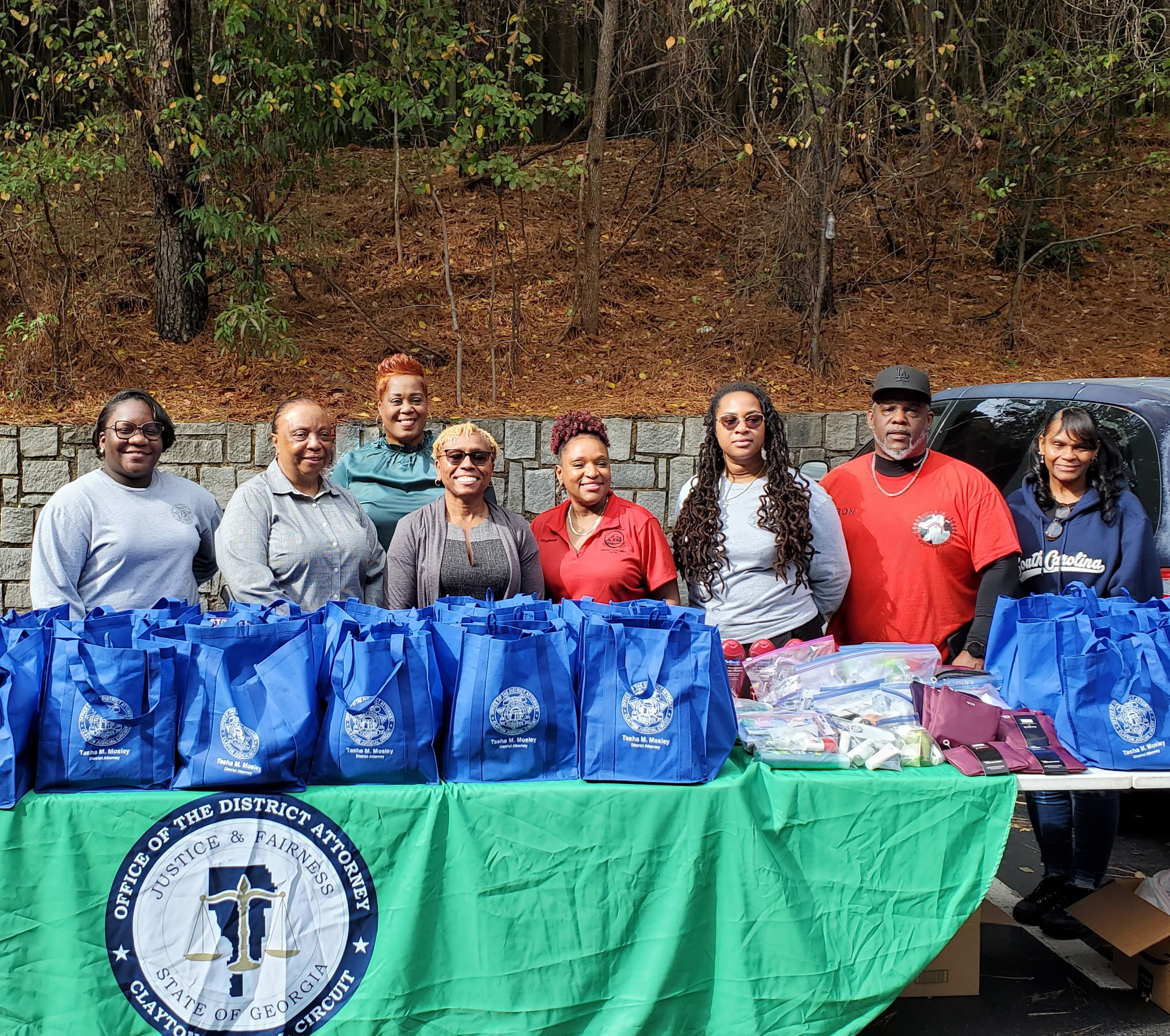 Group of eight people standing behind a table with blue bags and supplies outdoors, trees in background.