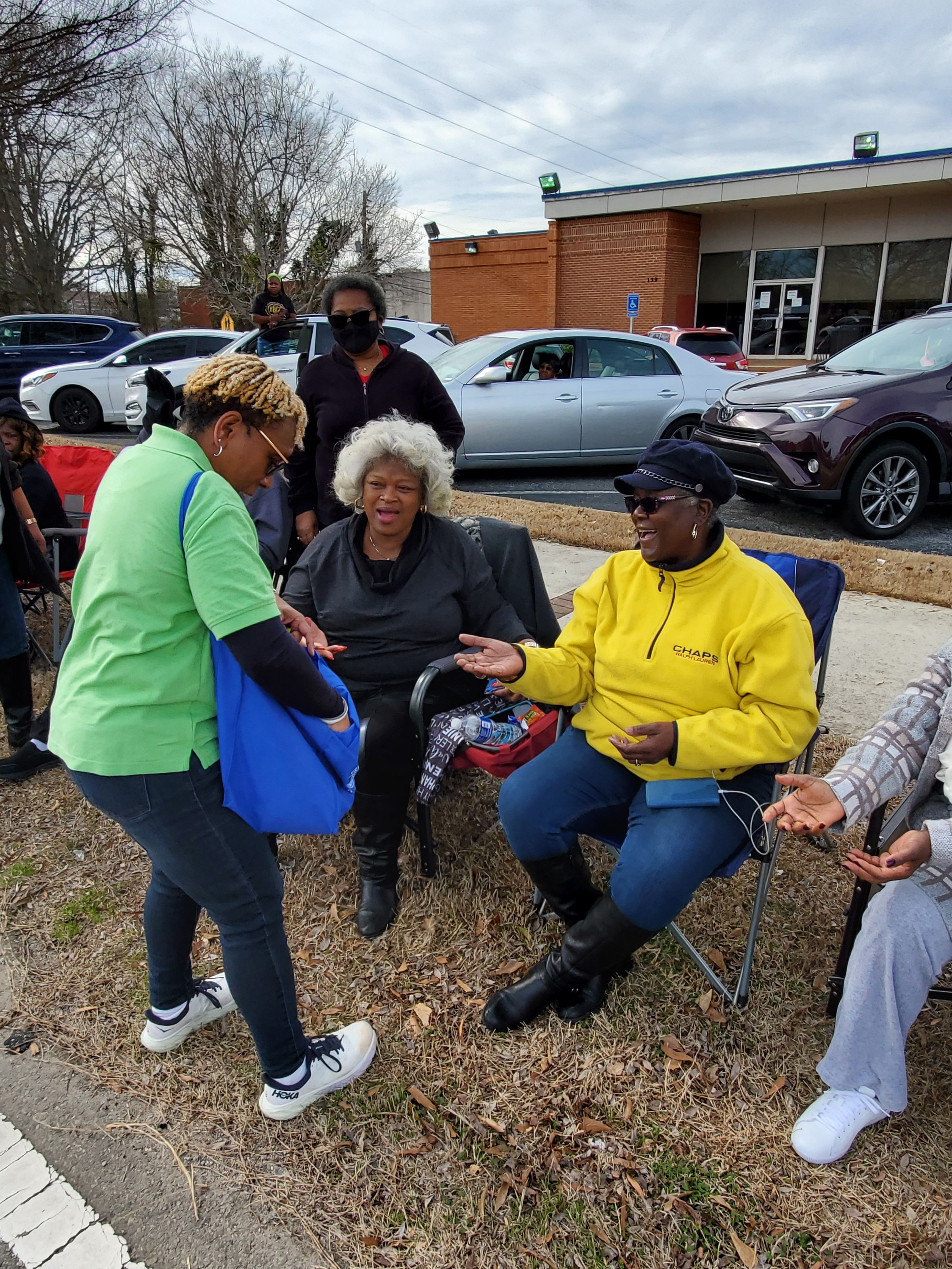 Four people sitting outdoors on a gravel surface with parked cars in the background, some holding drinks.
