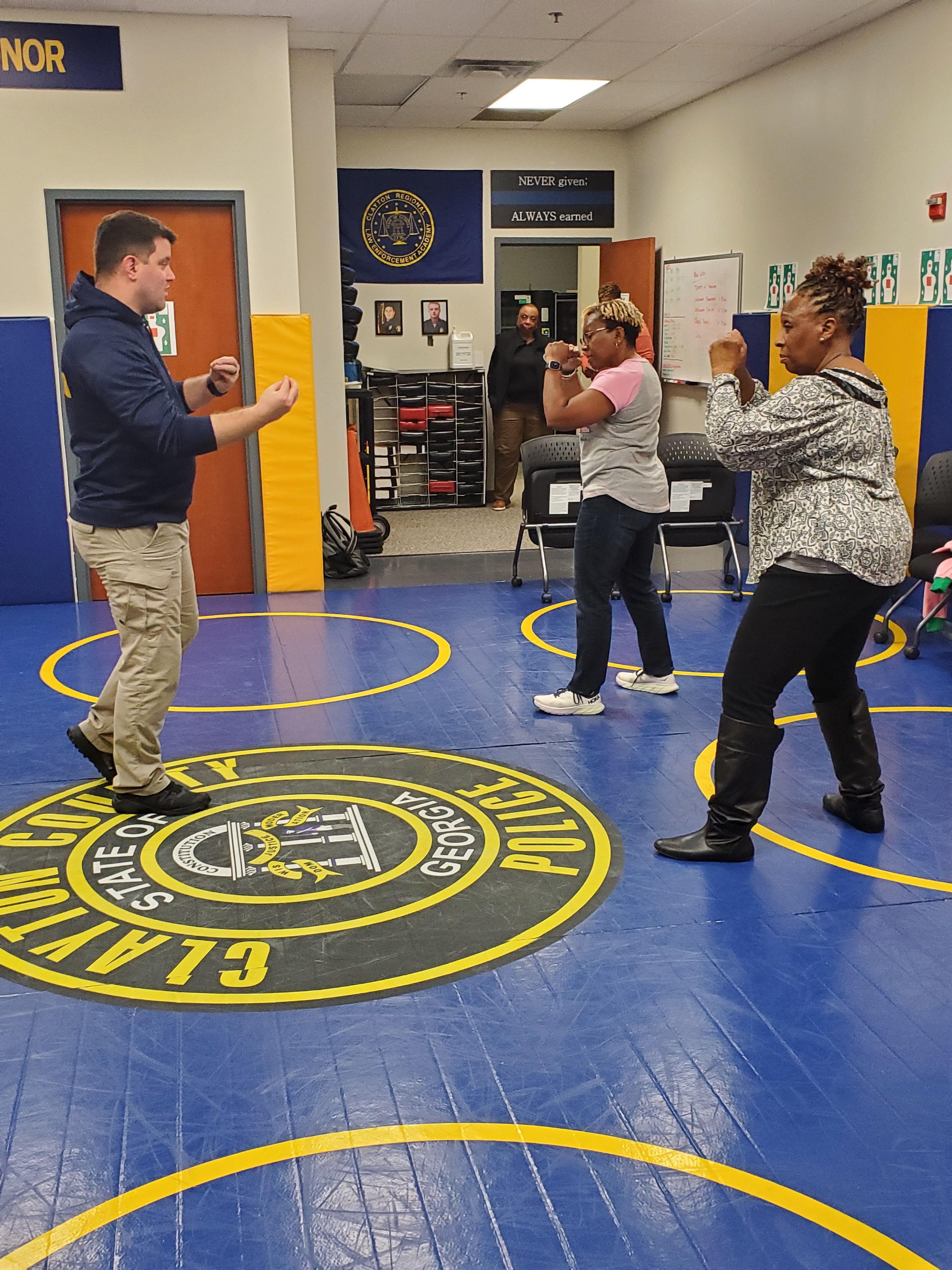 Two people standing on a blue wrestling mat with a yellow logo, in a room with posters and a vending machine.