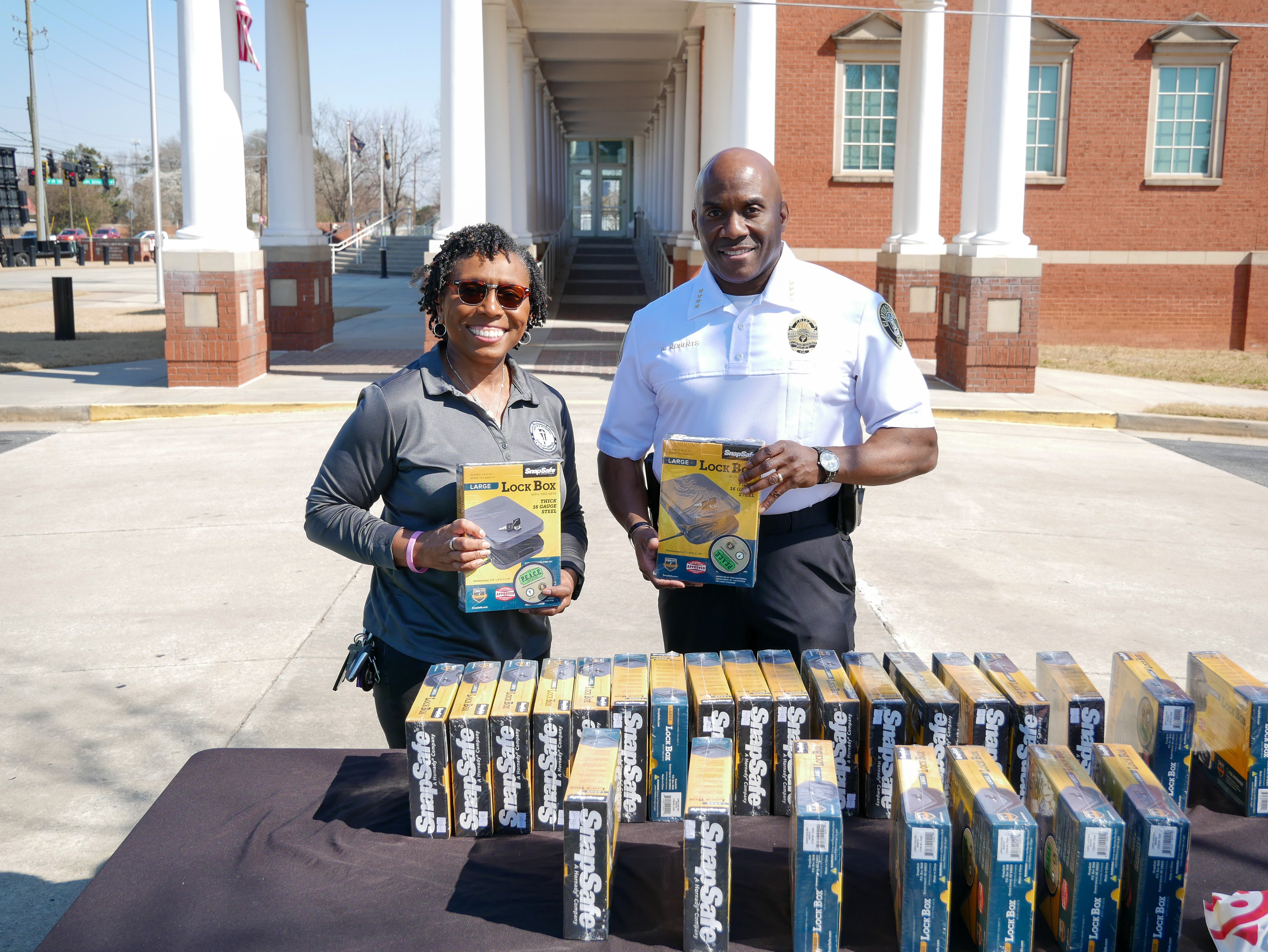 Two people stand outdoors holding brochures, with a table of brochures in front of them, in front of a building with columns.