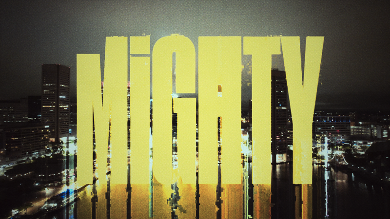 Bright yellow text spelling 'MIGHTY' over a cityscape at night, with reflections on water.