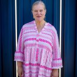 Older woman with gray hair in a pink and white patterned dress standing in front of a blue wall.