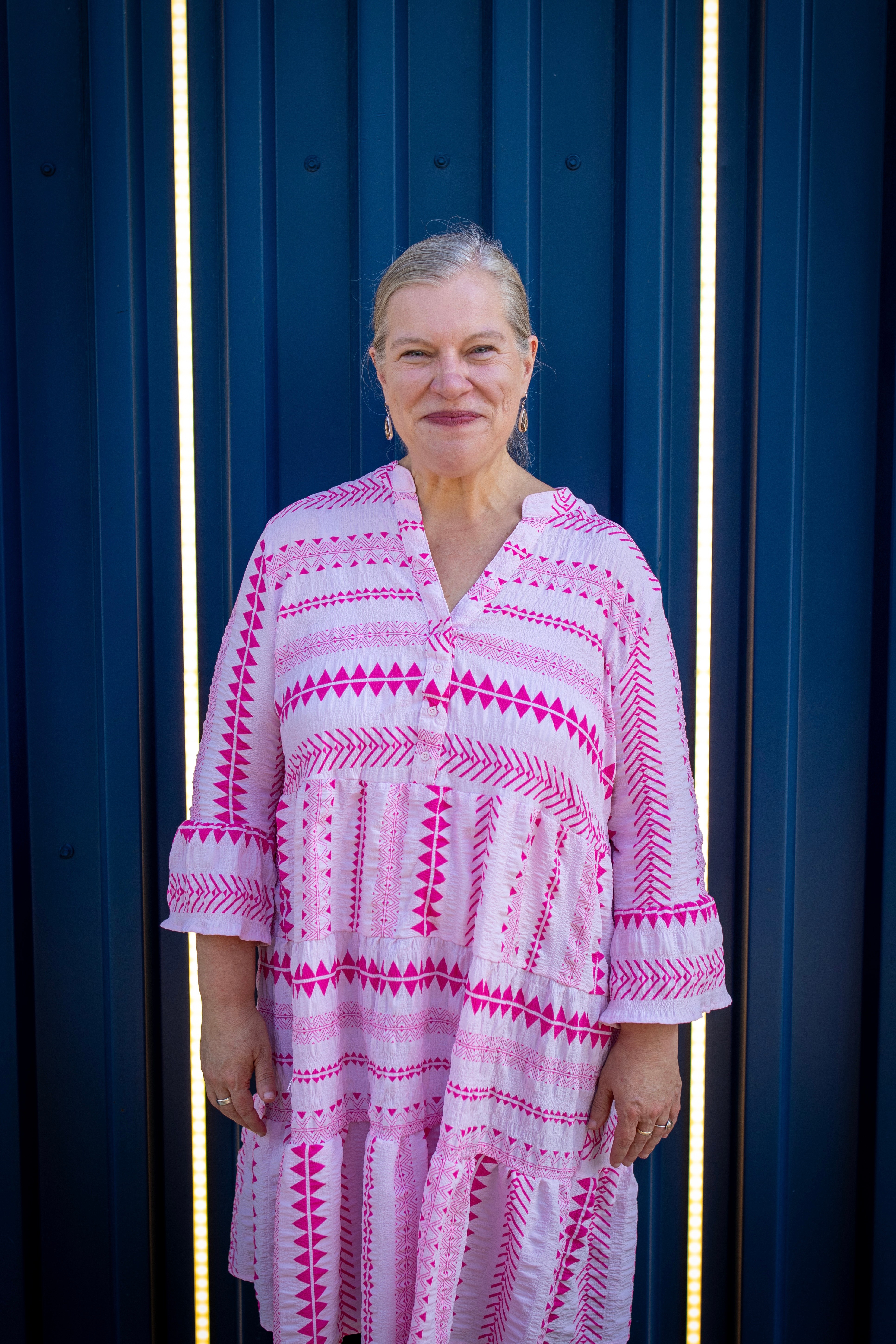 Older woman with gray hair in a pink and white patterned dress standing in front of a blue wall.