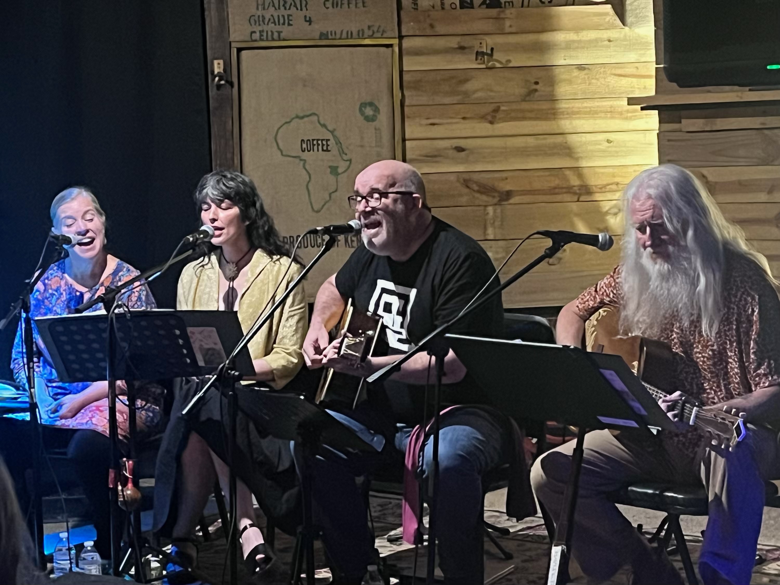 Four people sit on stage with microphones, singing and playing guitars, in a wooden-paneled setting.