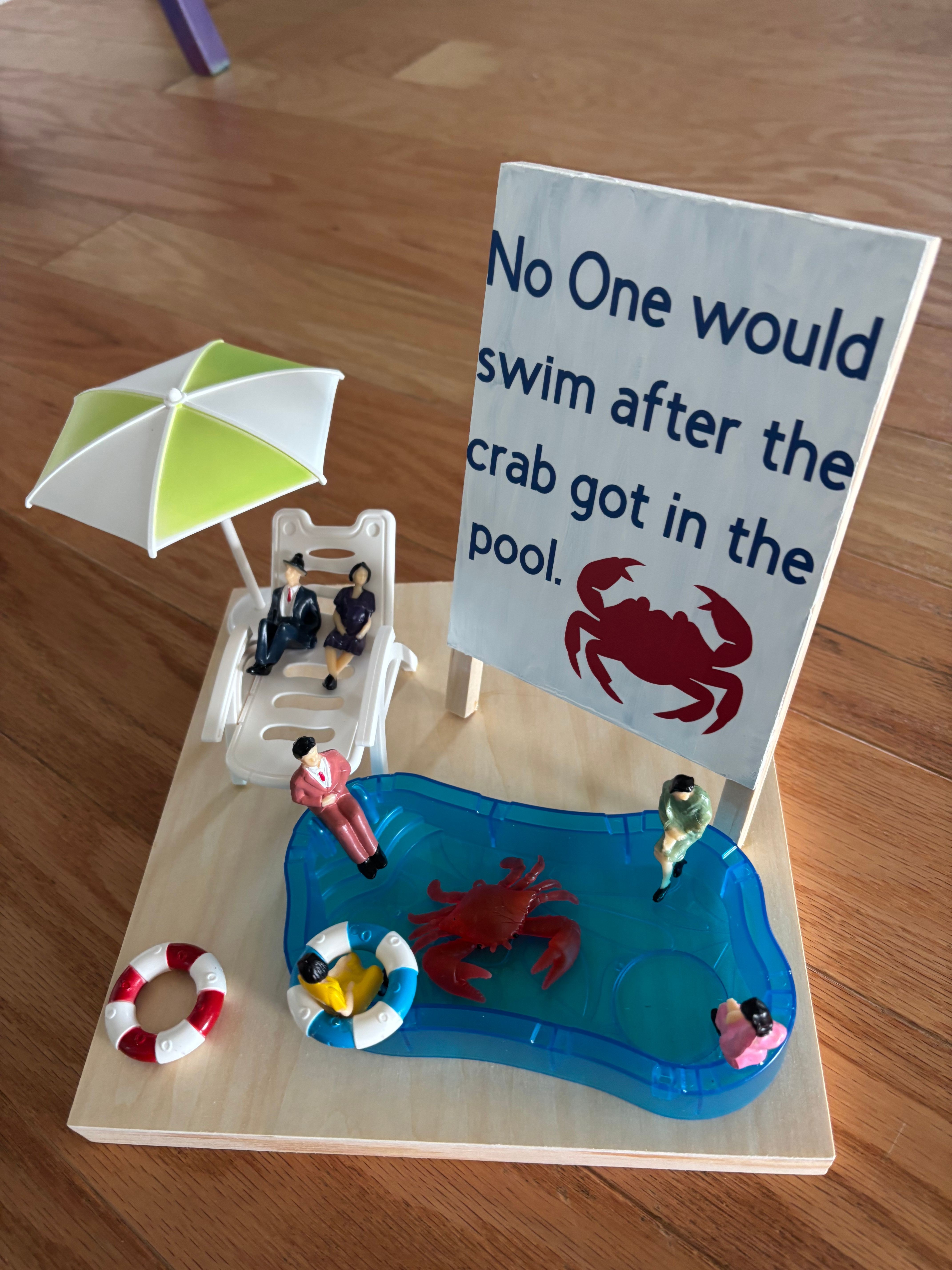 Miniature pool scene with a sign, umbrella, and small figures around a pool, on a wooden surface.