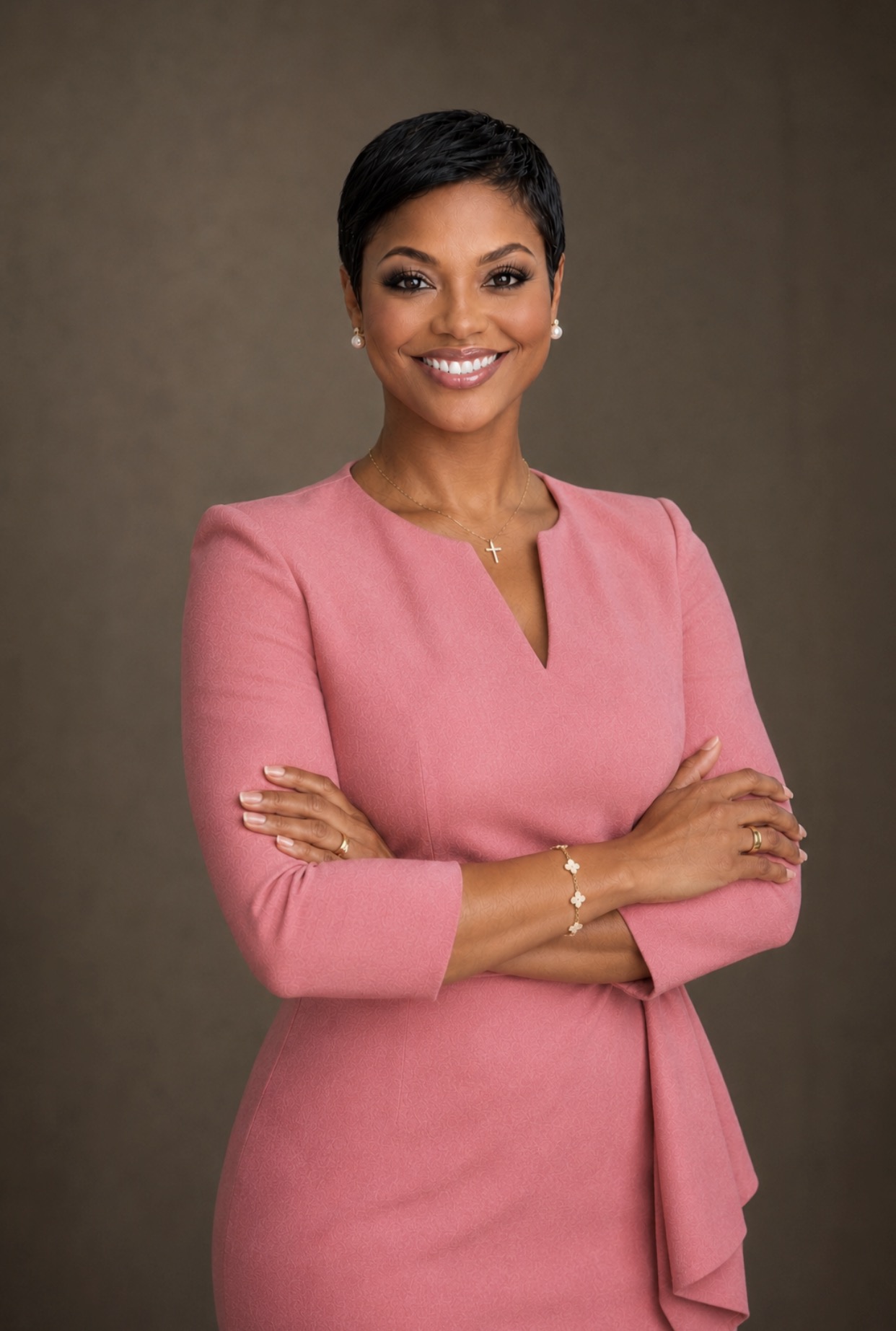 Smiling woman with short black hair wearing a pink dress with crossed arms, standing against a plain background.