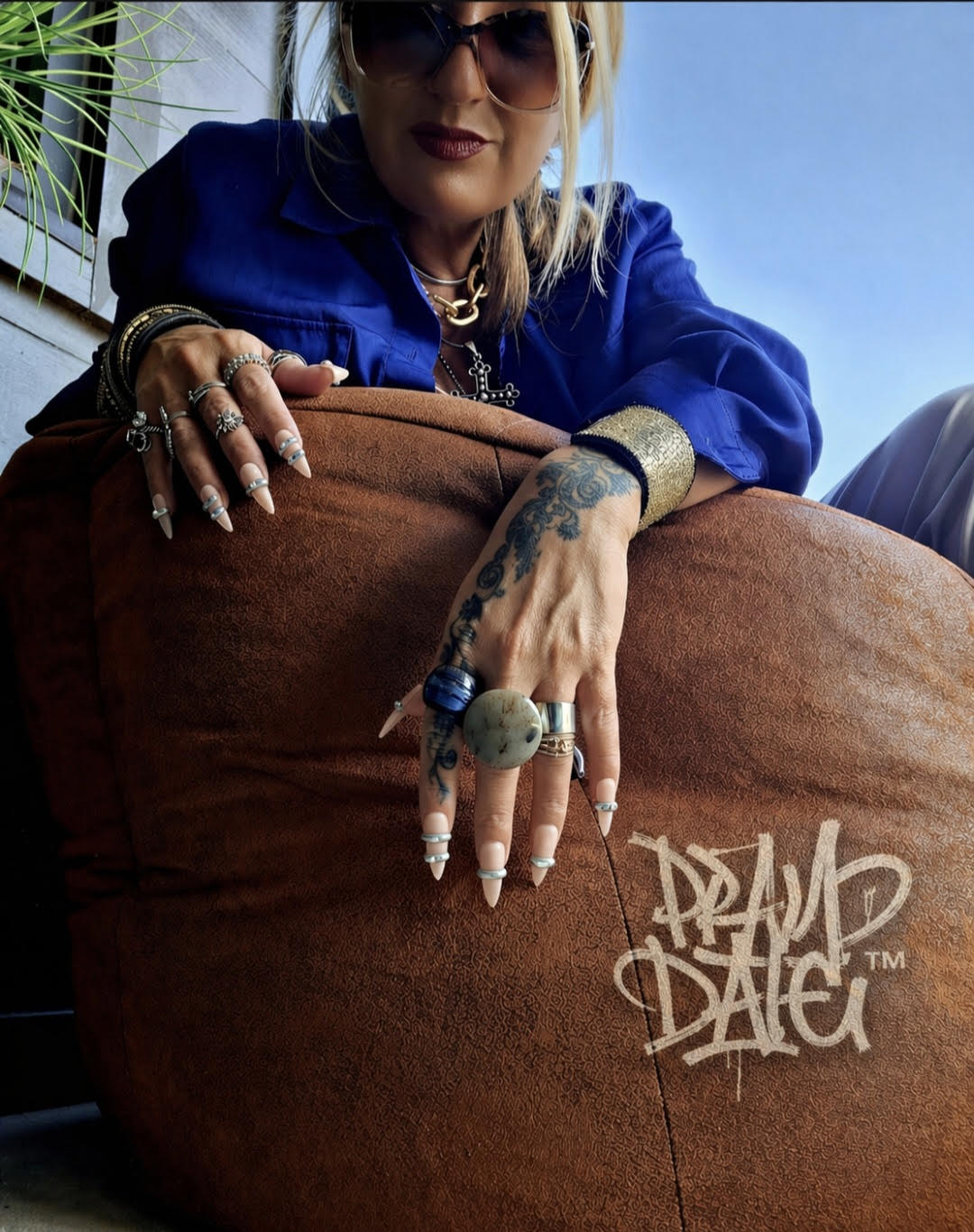 Person with blonde hair, sunglasses, and jewelry leans over a large brown cushion or bag, with a blue sky background.
