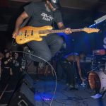 Person jumping on a stage playing a yellow electric bass guitar, wearing a black cap and t-shirt, with a band performing in the background.