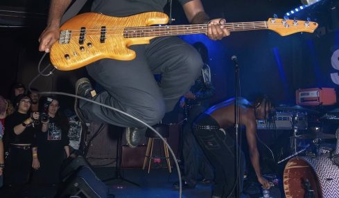 Person jumping on a stage playing a yellow electric bass guitar, wearing a black cap and t-shirt, with a band performing in the background.