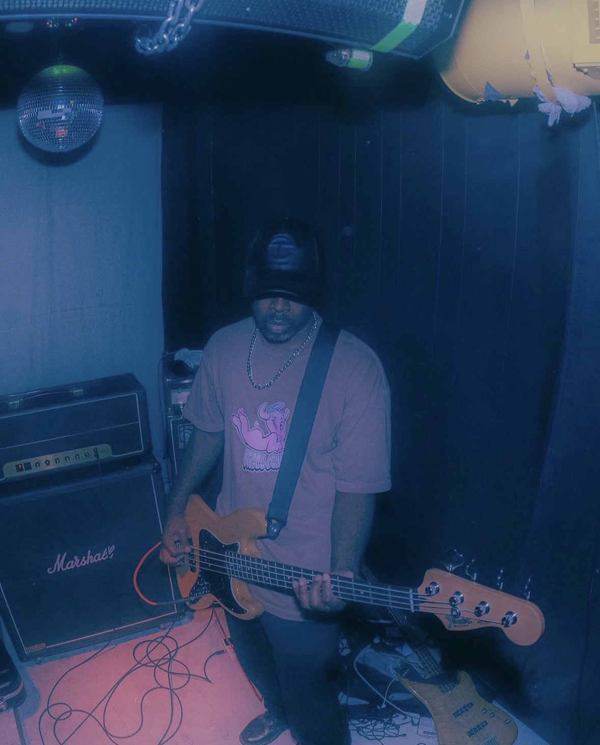 Person playing guitar in a dimly lit room with musical equipment, wearing a cap and glasses.
