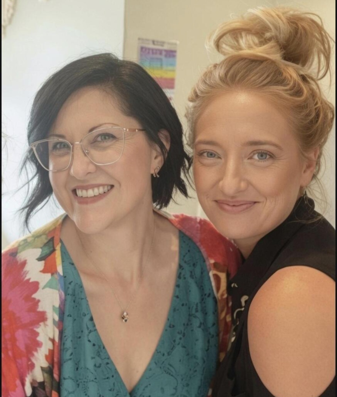 Two women smiling, one with glasses and dark hair, the other with blonde hair in a bun, in an indoor setting.