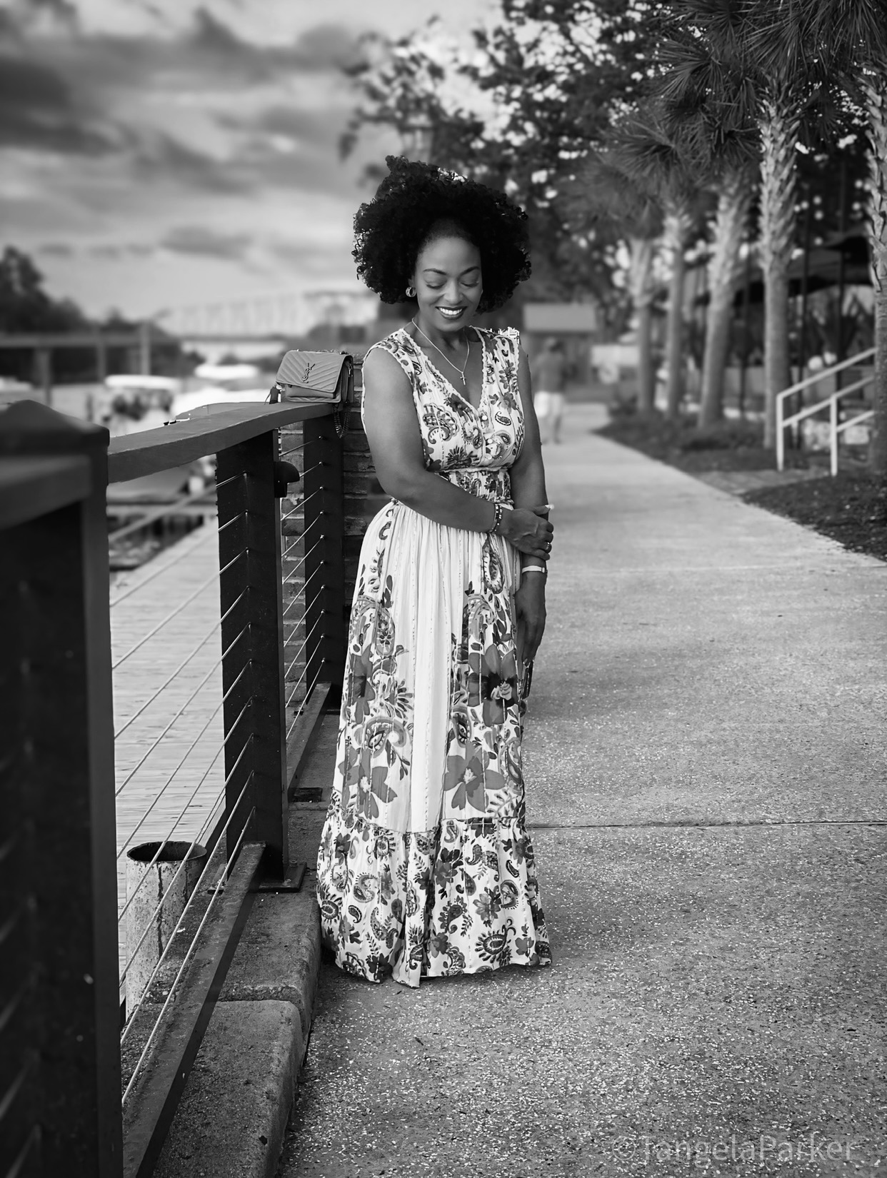 Woman with curly hair in a long patterned dress standing on a sidewalk near a railing, trees, and cloudy sky.