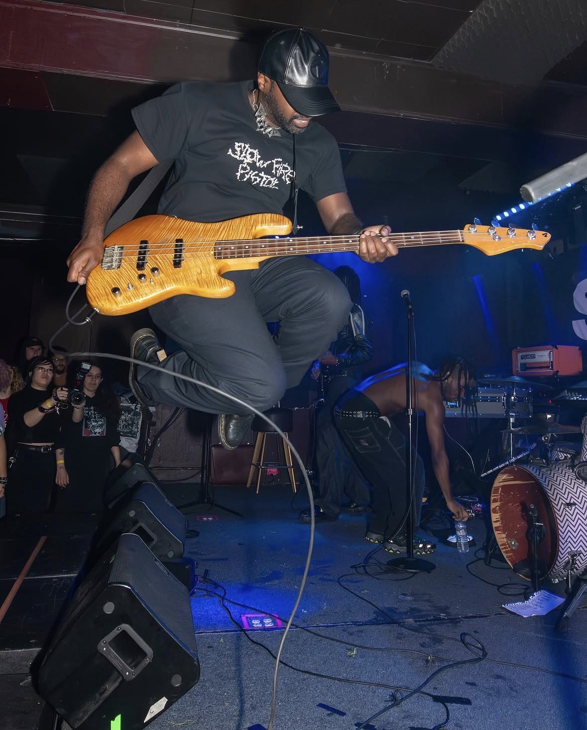 Person playing electric bass guitar on stage with audience in background, stage equipment visible.