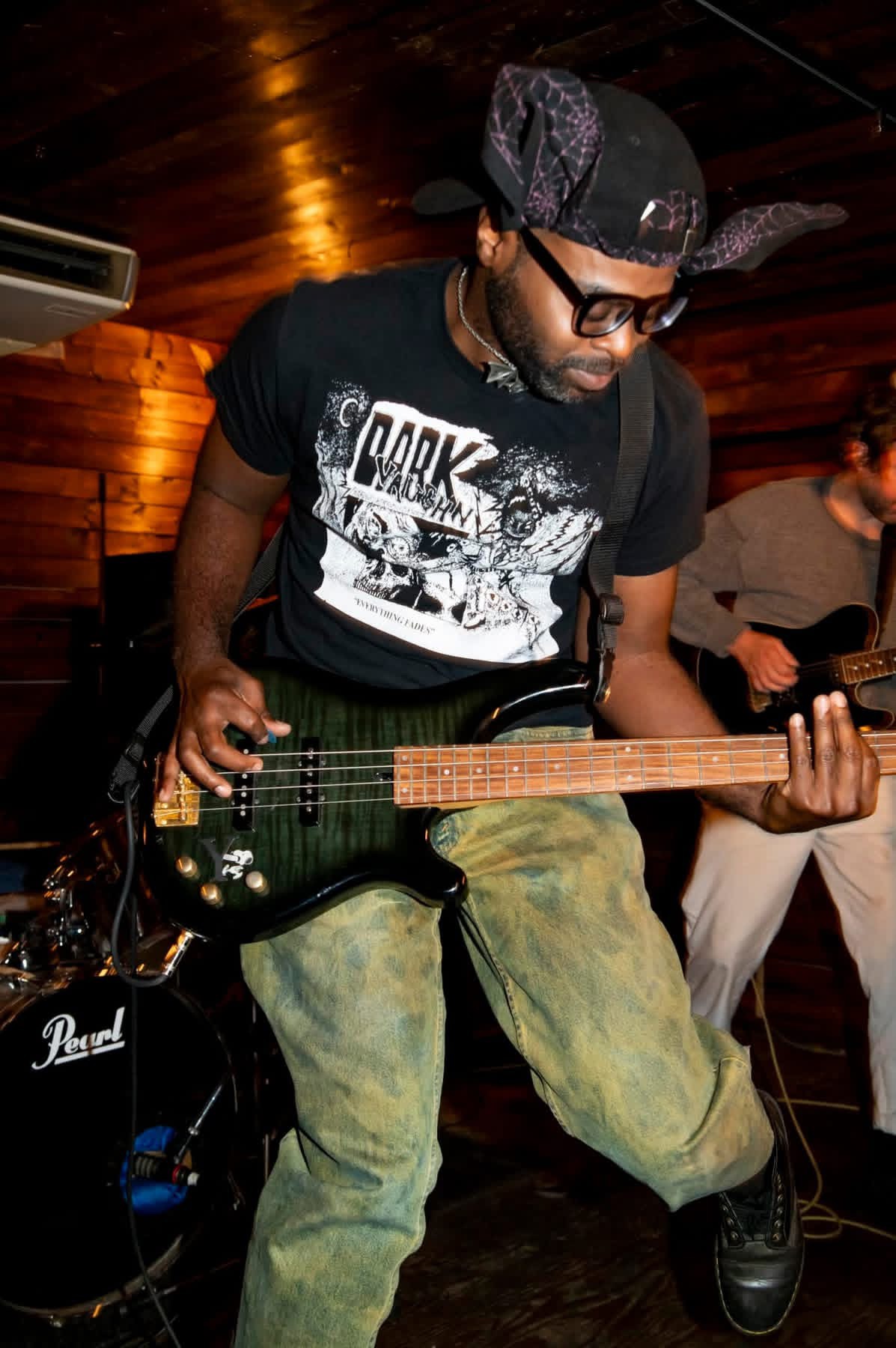 Person playing electric guitar on stage with wooden background, wearing glasses, a cap, and casual clothing.
