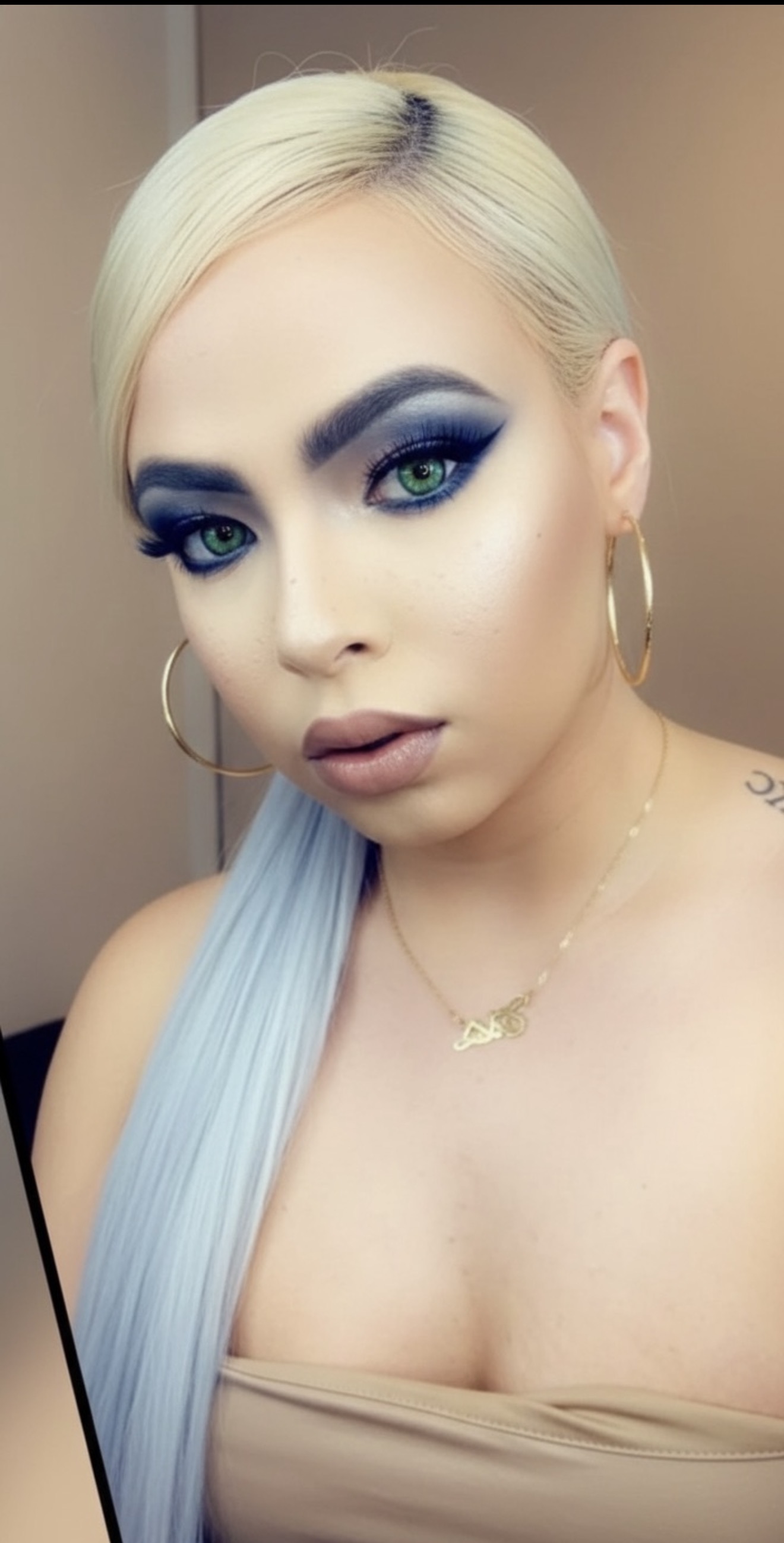 Close-up of a woman with platinum blonde hair, bold eye makeup, hoop earrings, and a light blue scarf.