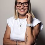 Woman with glasses smiling, wearing a white top, standing against a plain background.