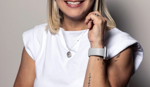 Woman with glasses smiling, wearing a white top, standing against a plain background.