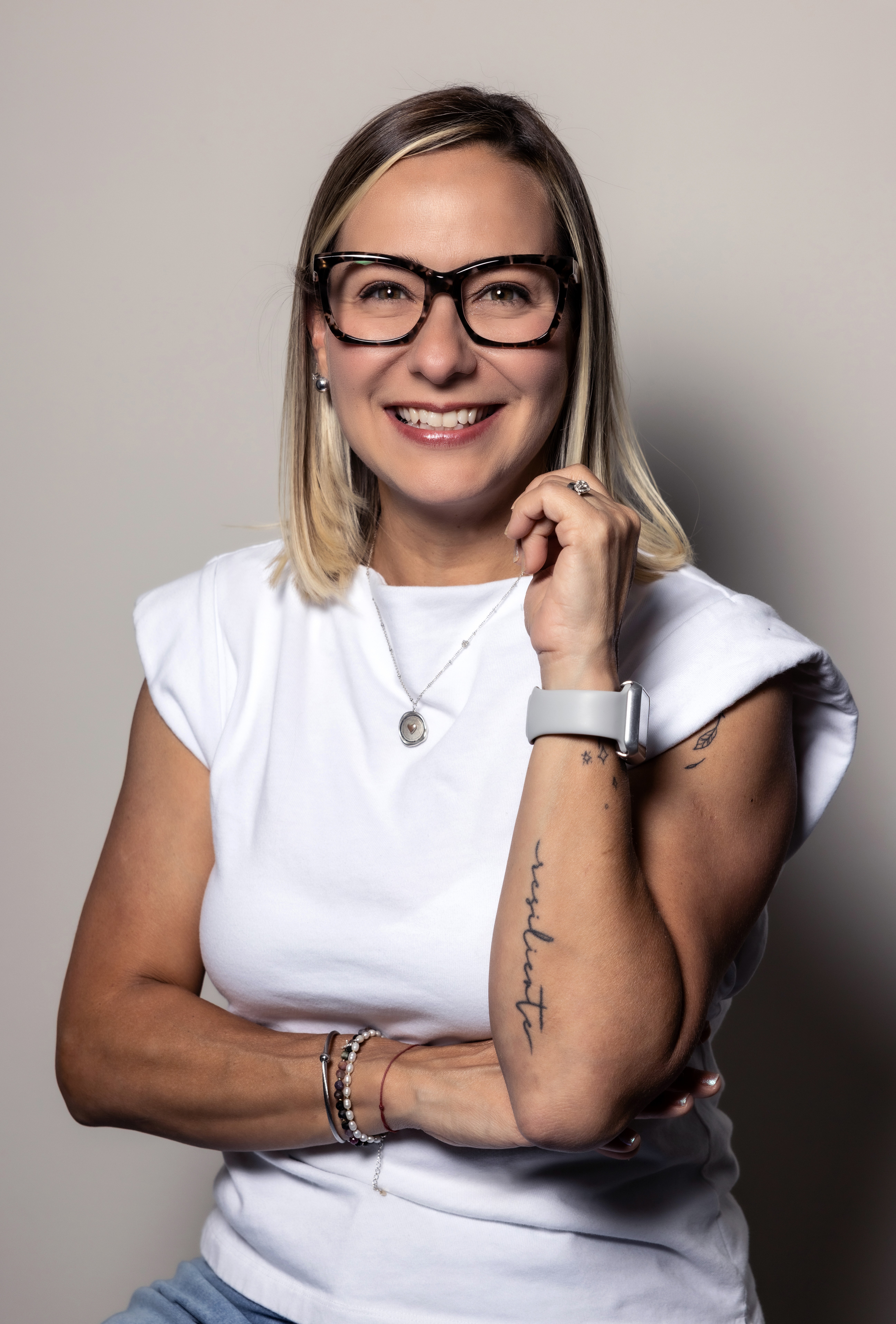 Woman with glasses smiling, wearing a white top, standing against a plain background.