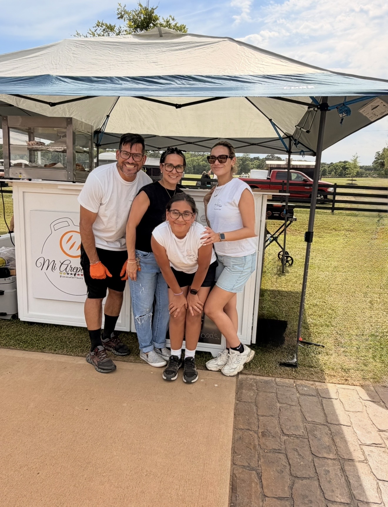 Four people standing under a canopy outdoors, smiling, with a grassy field and fence in background.