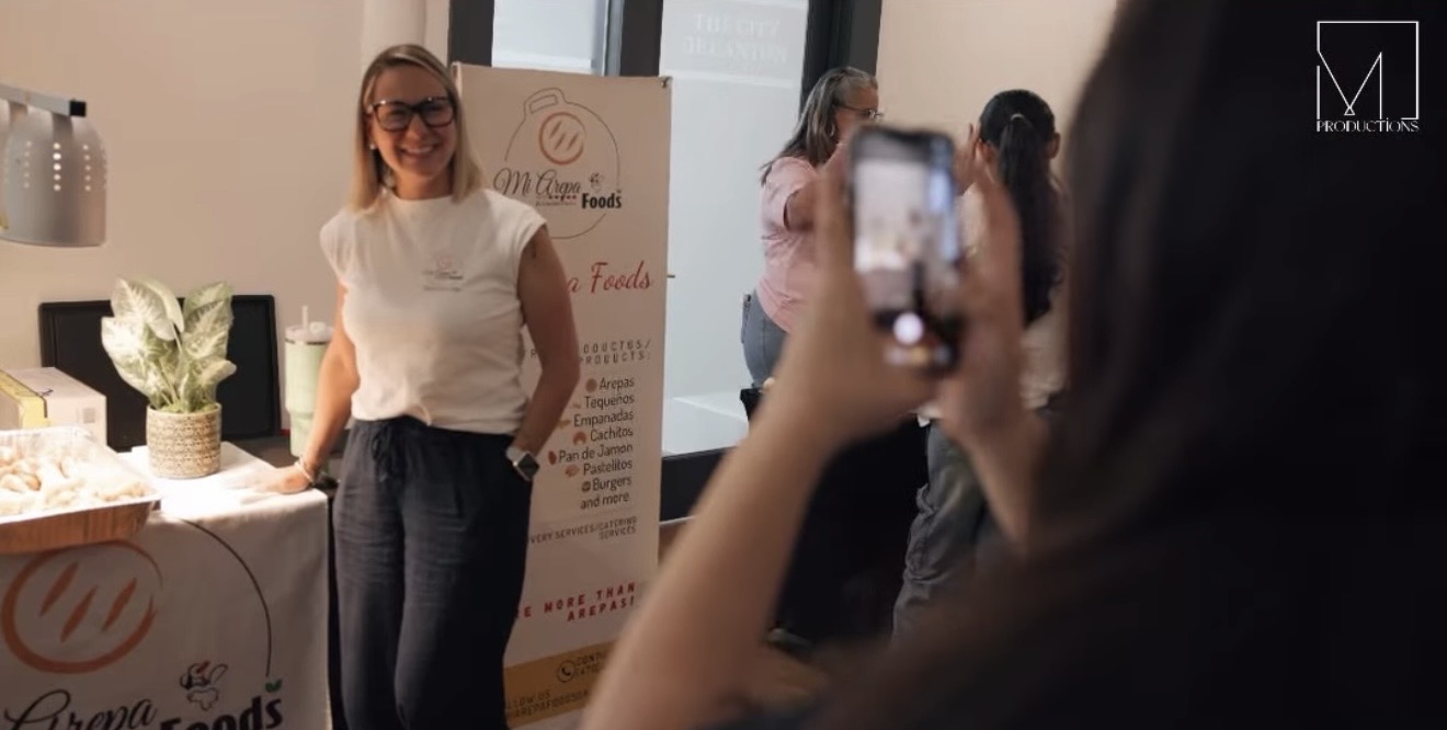 Woman with glasses smiling at camera, standing near a table with a plant and promotional banner, people in background.