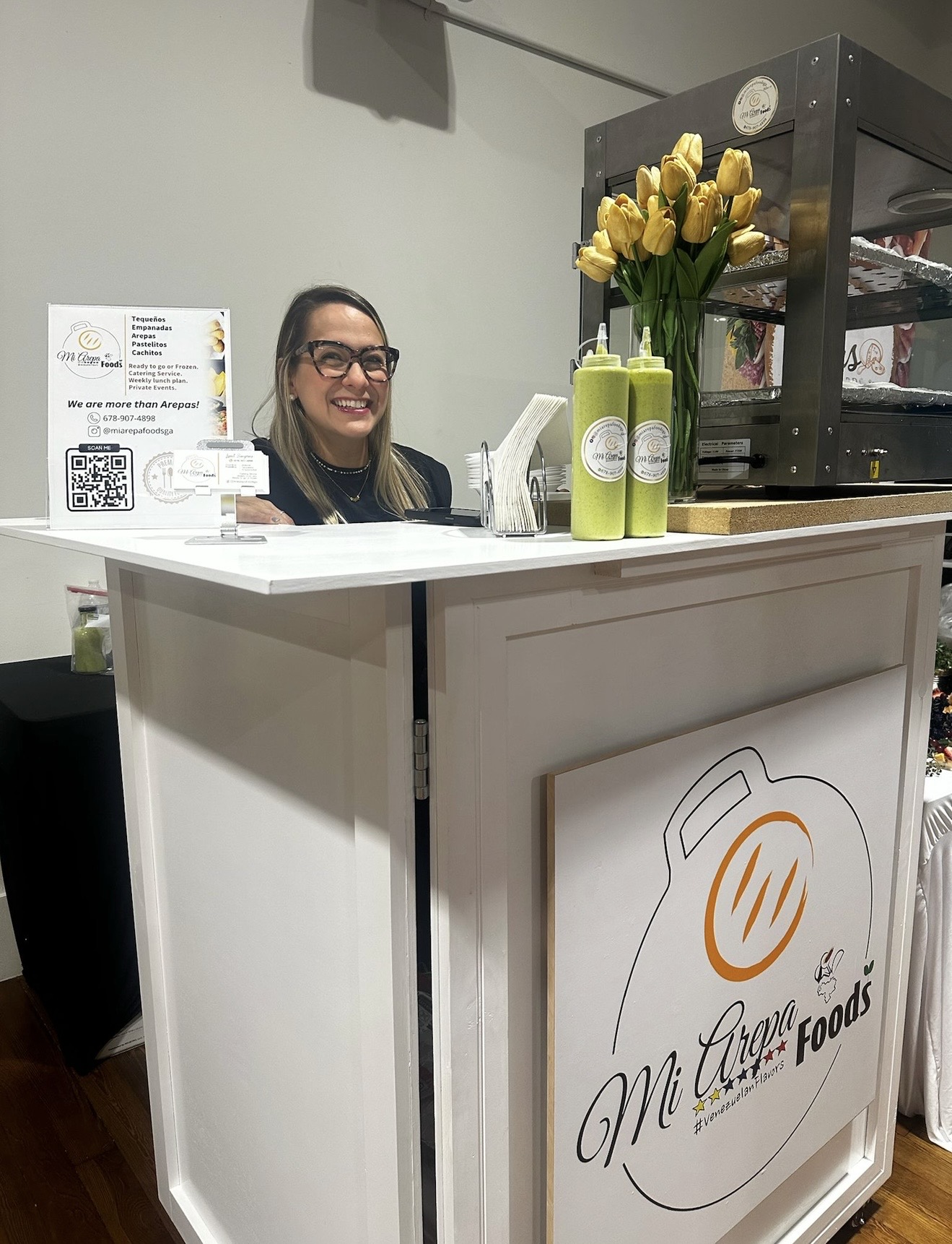Smiling woman behind a white counter with yellow flowers and a coffee machine, in a cafe setting.