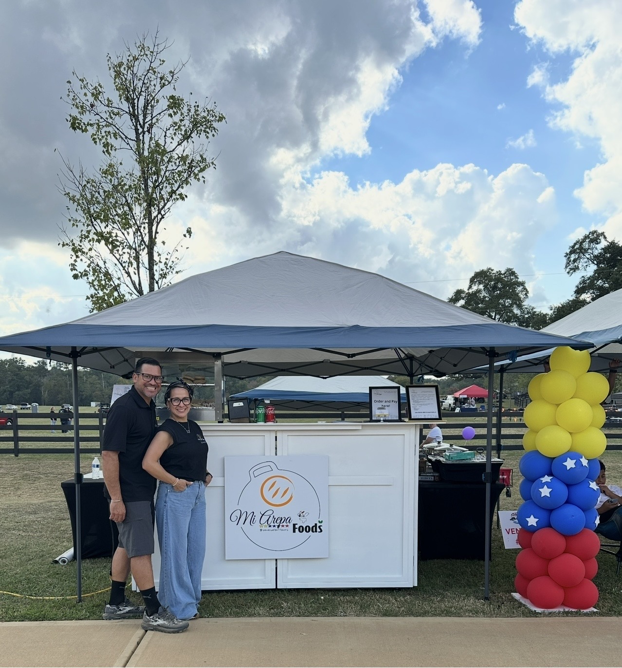 Two people standing in front of a food stand under a canopy, with colorful balloon decorations on the right, outdoors on a cloudy day.