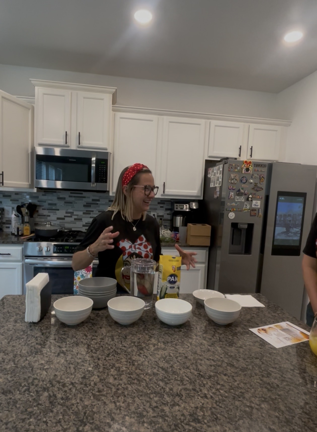 Woman with glasses and red headband preparing food in a kitchen with white cabinets and a refrigerator covered in magnets.