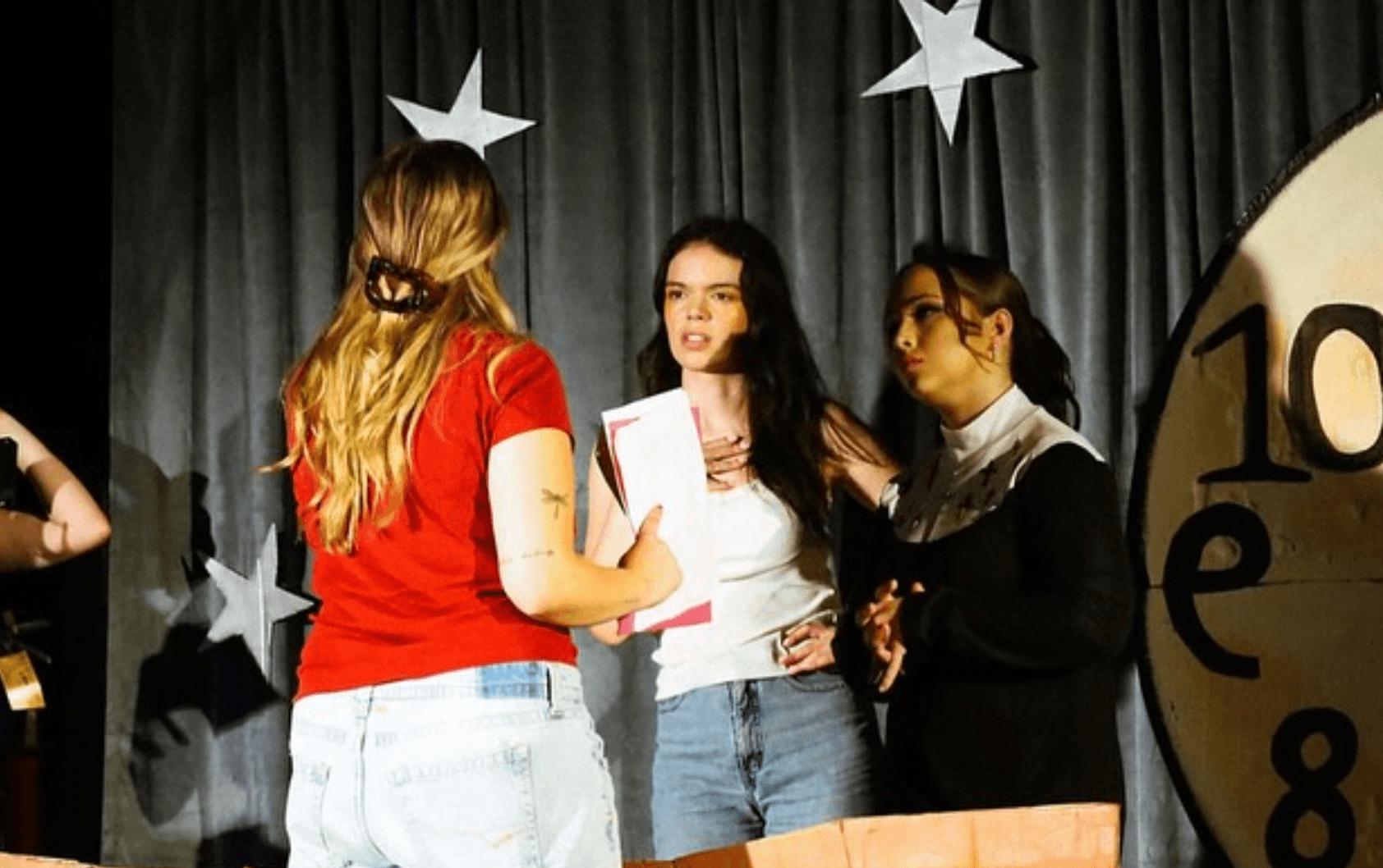 Three young women standing and talking on a stage with star decorations and a large clock face in the background.