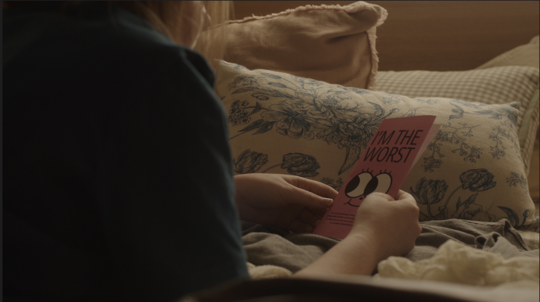 Person reading a pink book titled 'At the Wors' on a bed with patterned pillows.