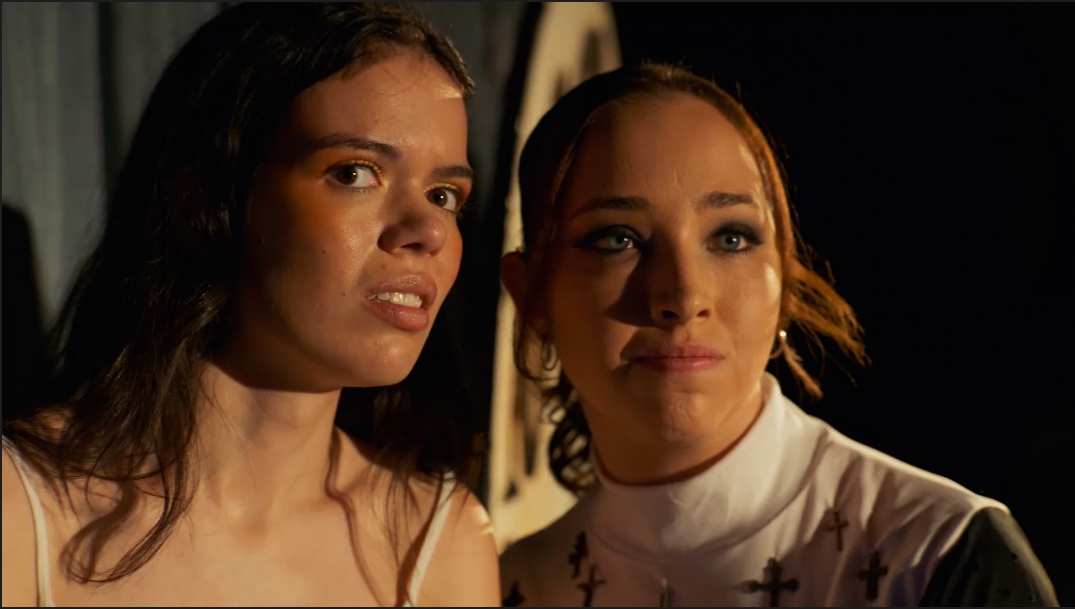 Two women with long hair, one with dark hair and the other with light hair, standing close together in a dimly lit setting.