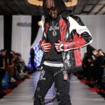 Male model walking on runway wearing a black, red, and white leather jacket, black pants, and sunglasses, with audience in background.