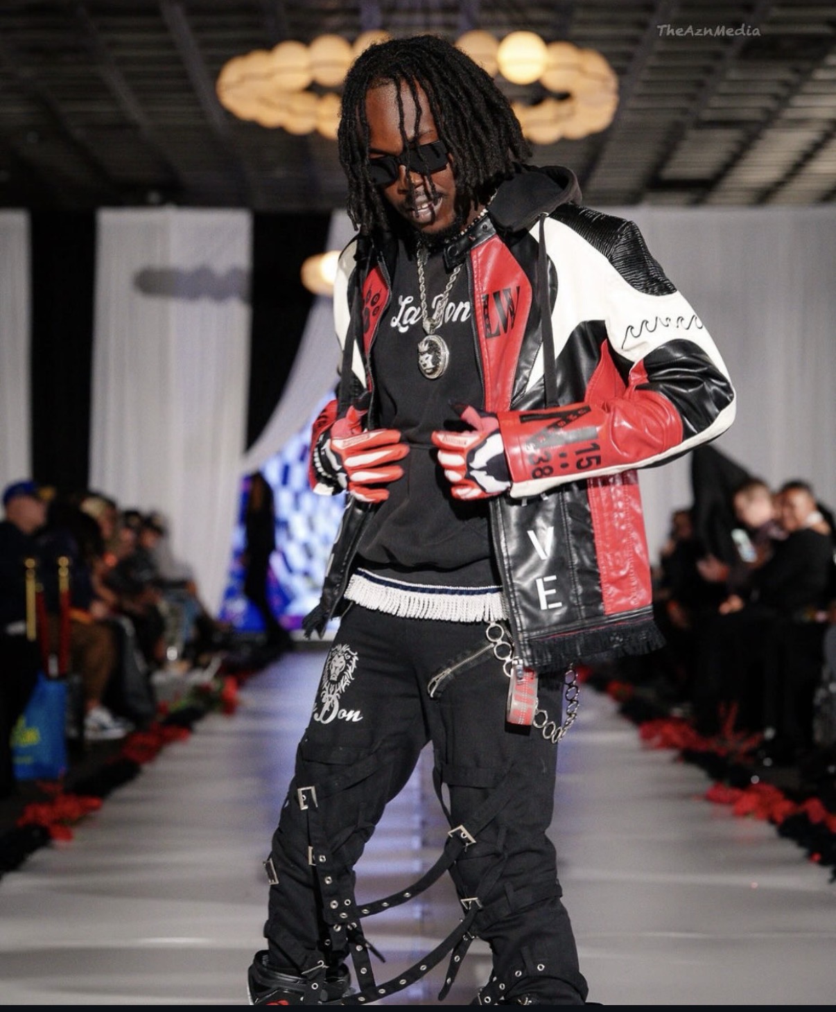 Male model walking on runway wearing a black, red, and white leather jacket, black pants, and sunglasses, with audience in background.