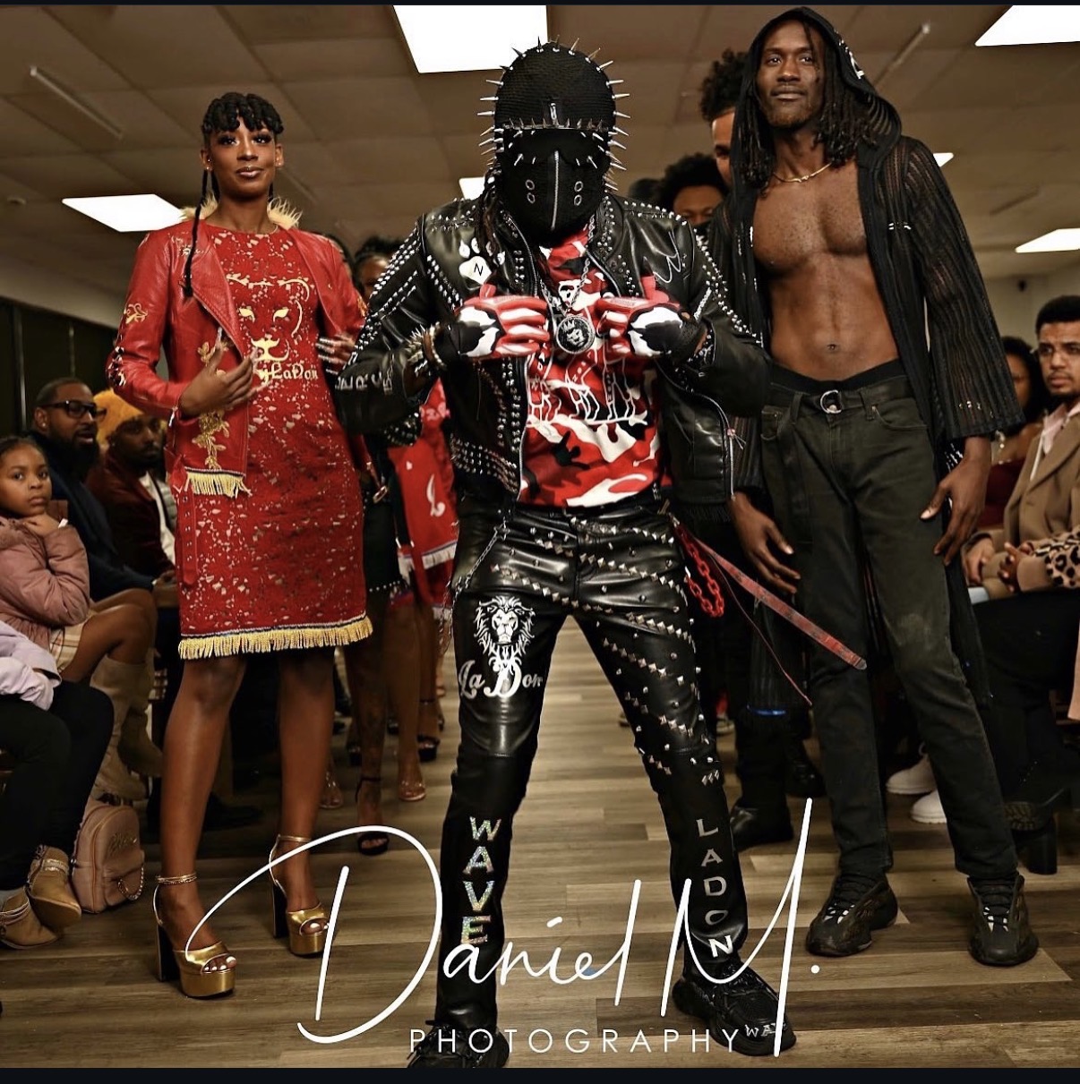 Three people standing in a room with an audience, one in a red dress, one in black leather with spikes, and one shirtless with a jacket.