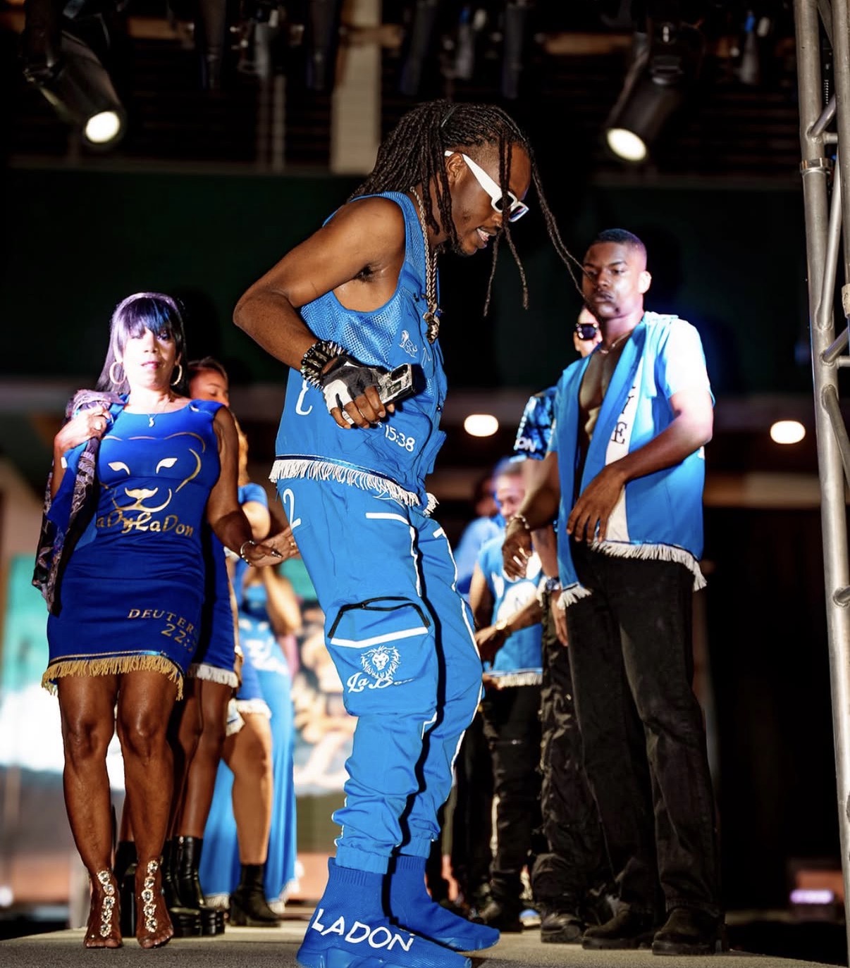 Group of people on stage, one person with dreadlocks and sunglasses, others in blue outfits, stage lighting overhead.