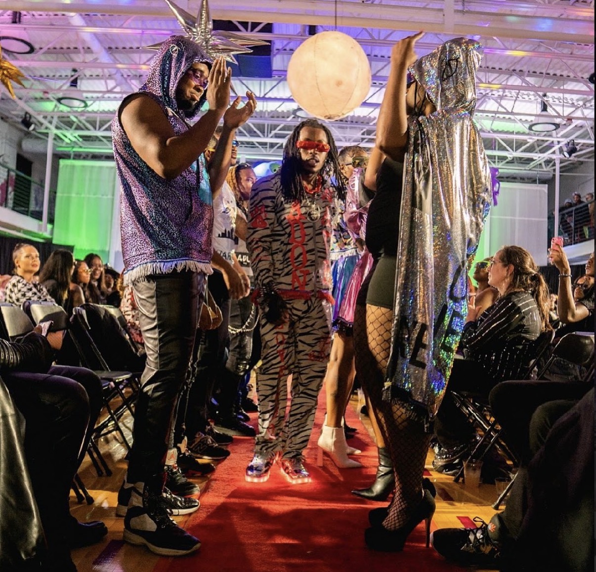 People walking on a red carpet at an indoor event with colorful lighting and a large moon decoration overhead.