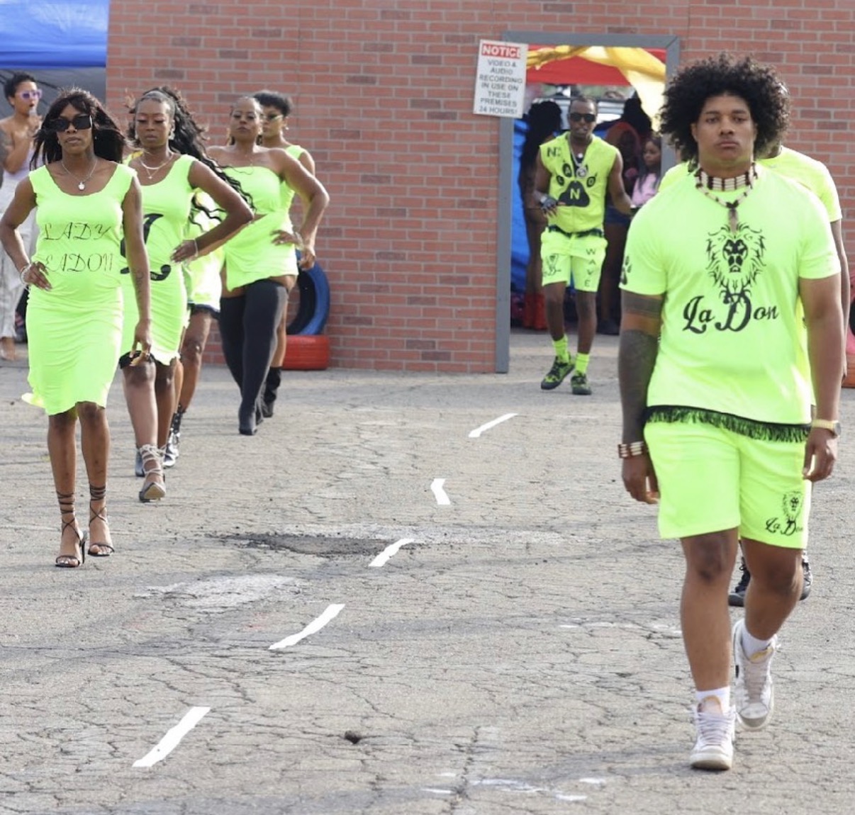 Group of people walking outdoors, some wearing bright green outfits, brick wall in background, pavement with white lines.