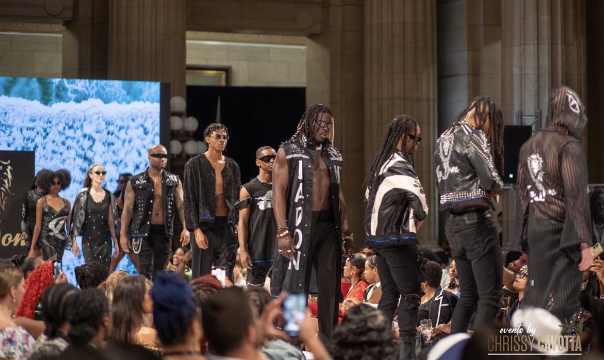 Group of models walking on runway during fashion show in large indoor venue with audience watching.
