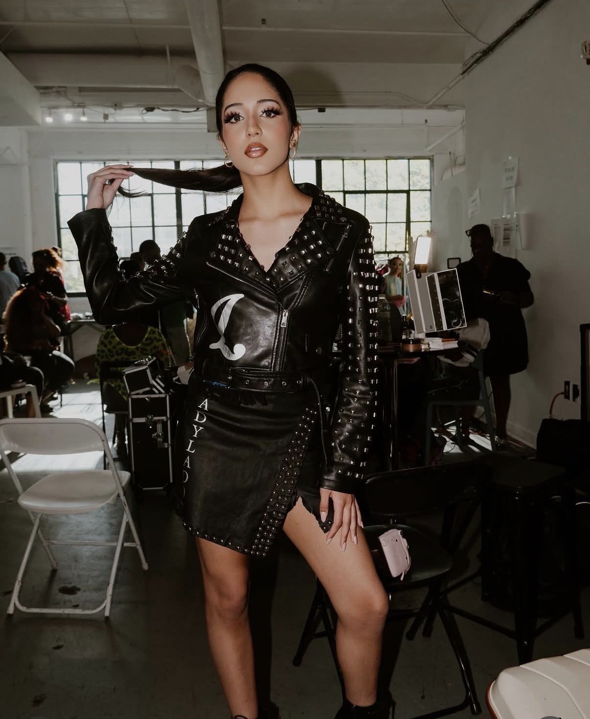 Woman with long braid, wearing black leather jacket and skirt, standing in a room with people and equipment in background.