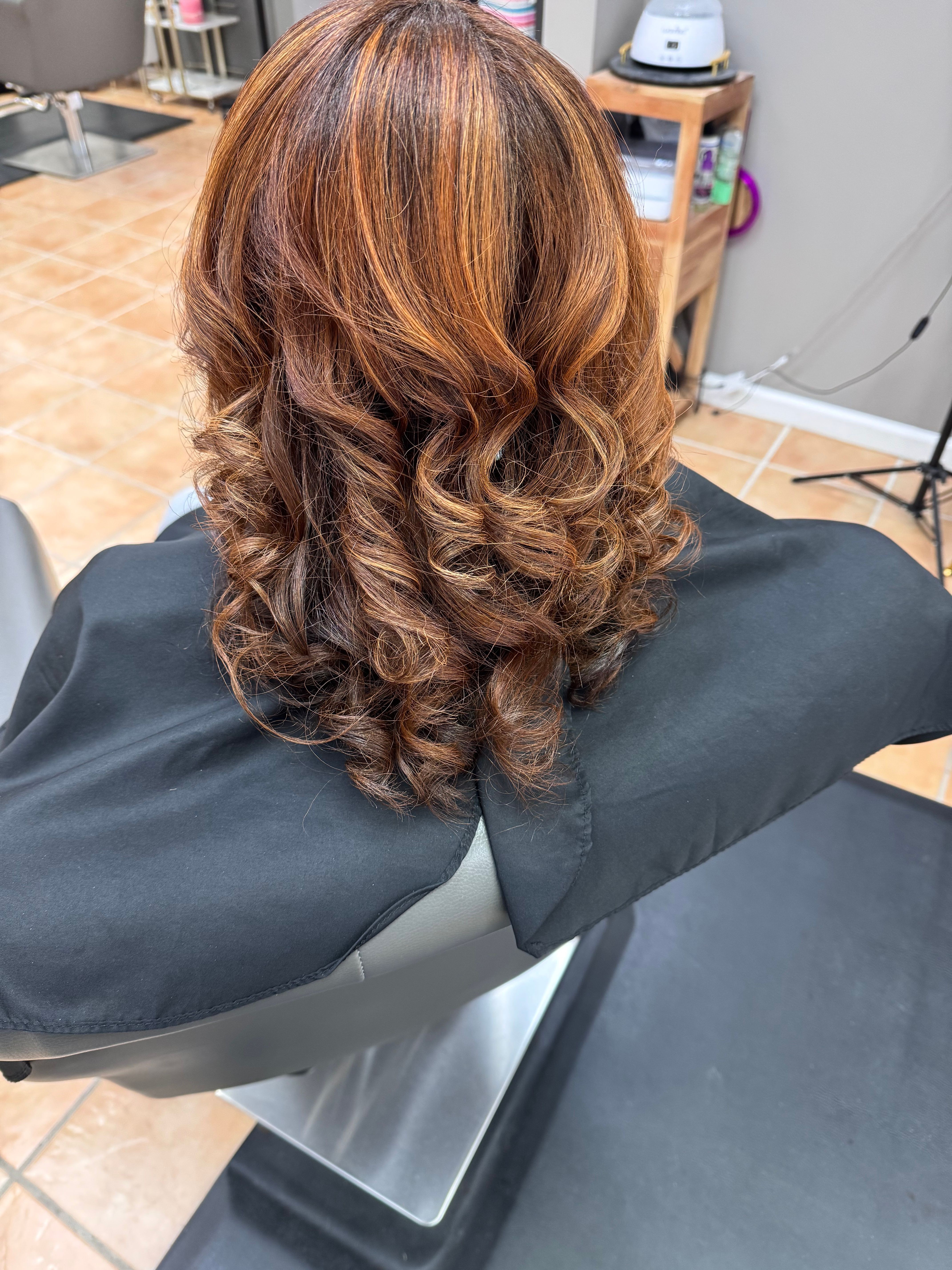 Person with curly, brown hair sitting in a chair, wearing a black cape, in a salon or similar setting.