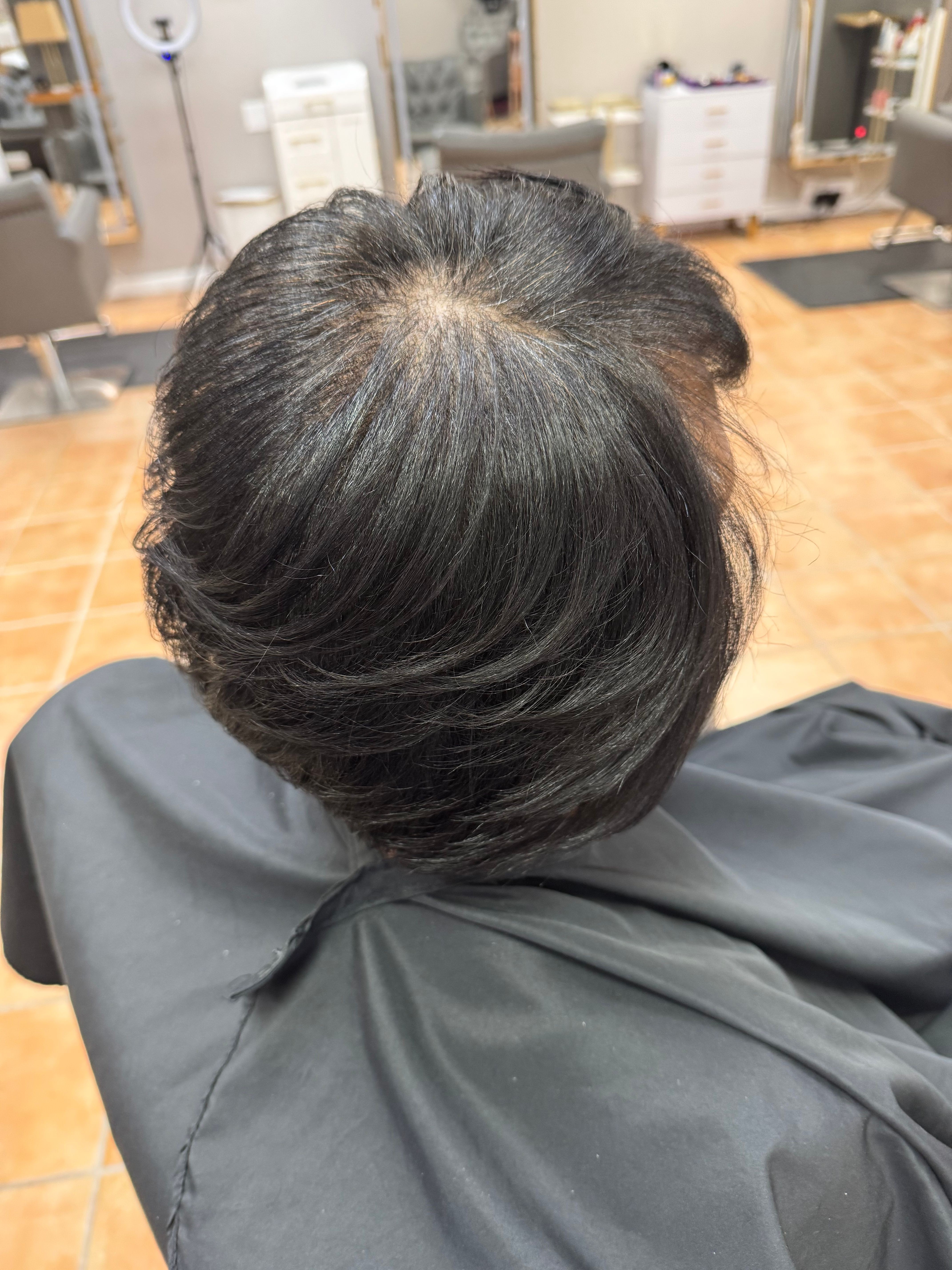Top view of a person's head with dark hair, showing a grayish scalp area, in a salon setting.
