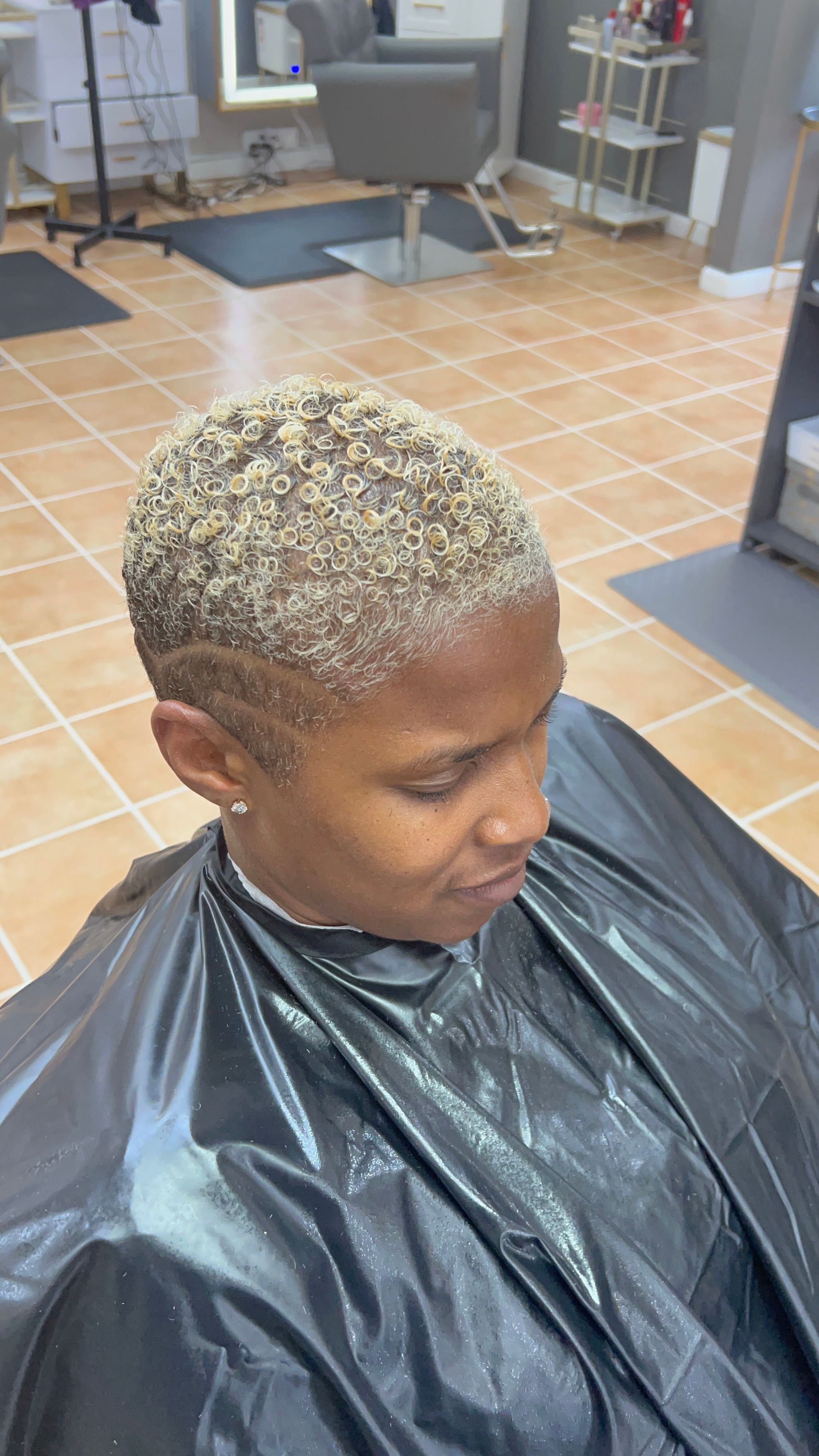 Person with short, curly, bleached hair sitting in a salon chair, wearing a black cape, in a salon setting.
