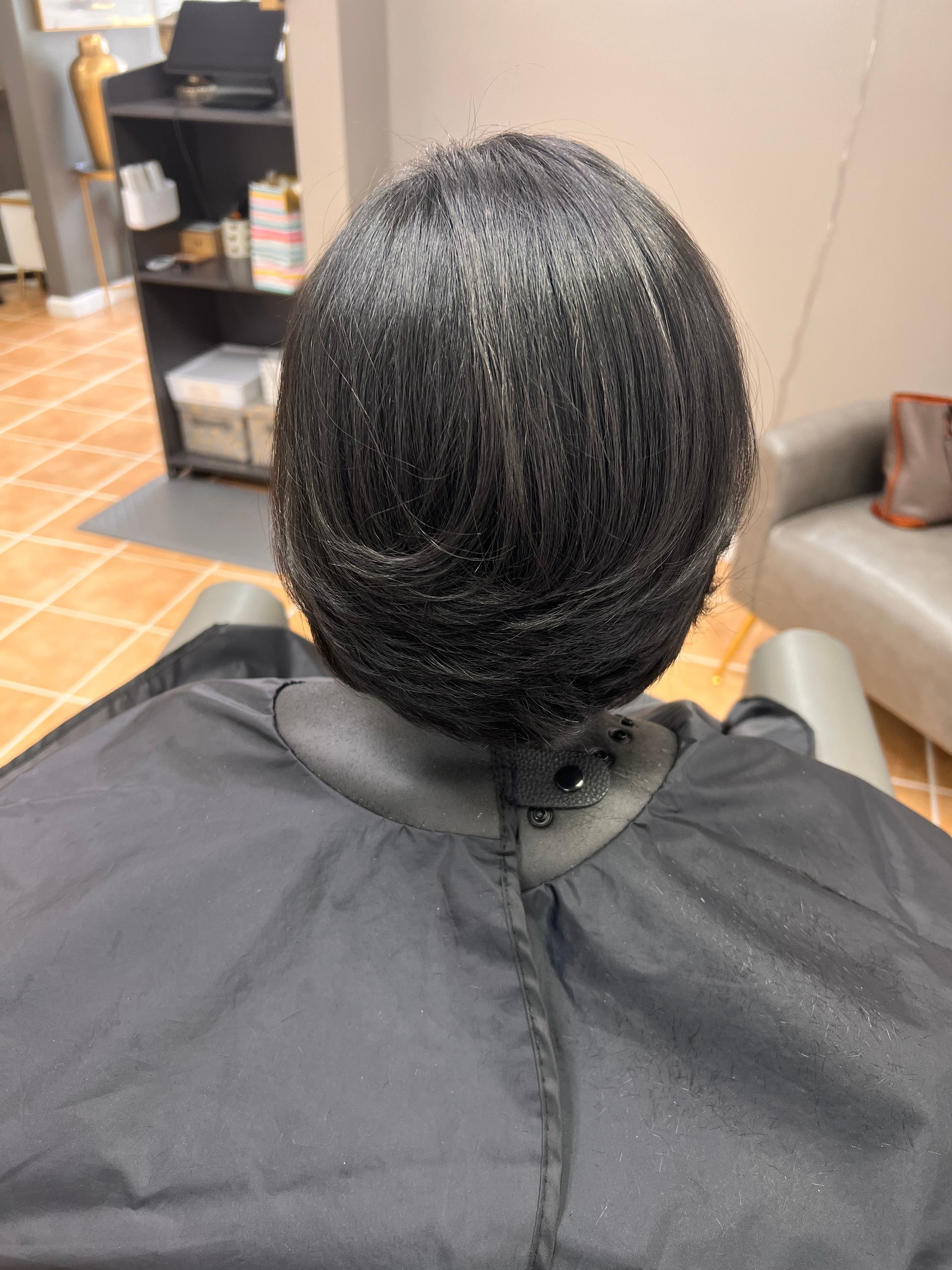 Person with dark hair sitting in a salon chair, facing away, with a black cape over their shoulders.
