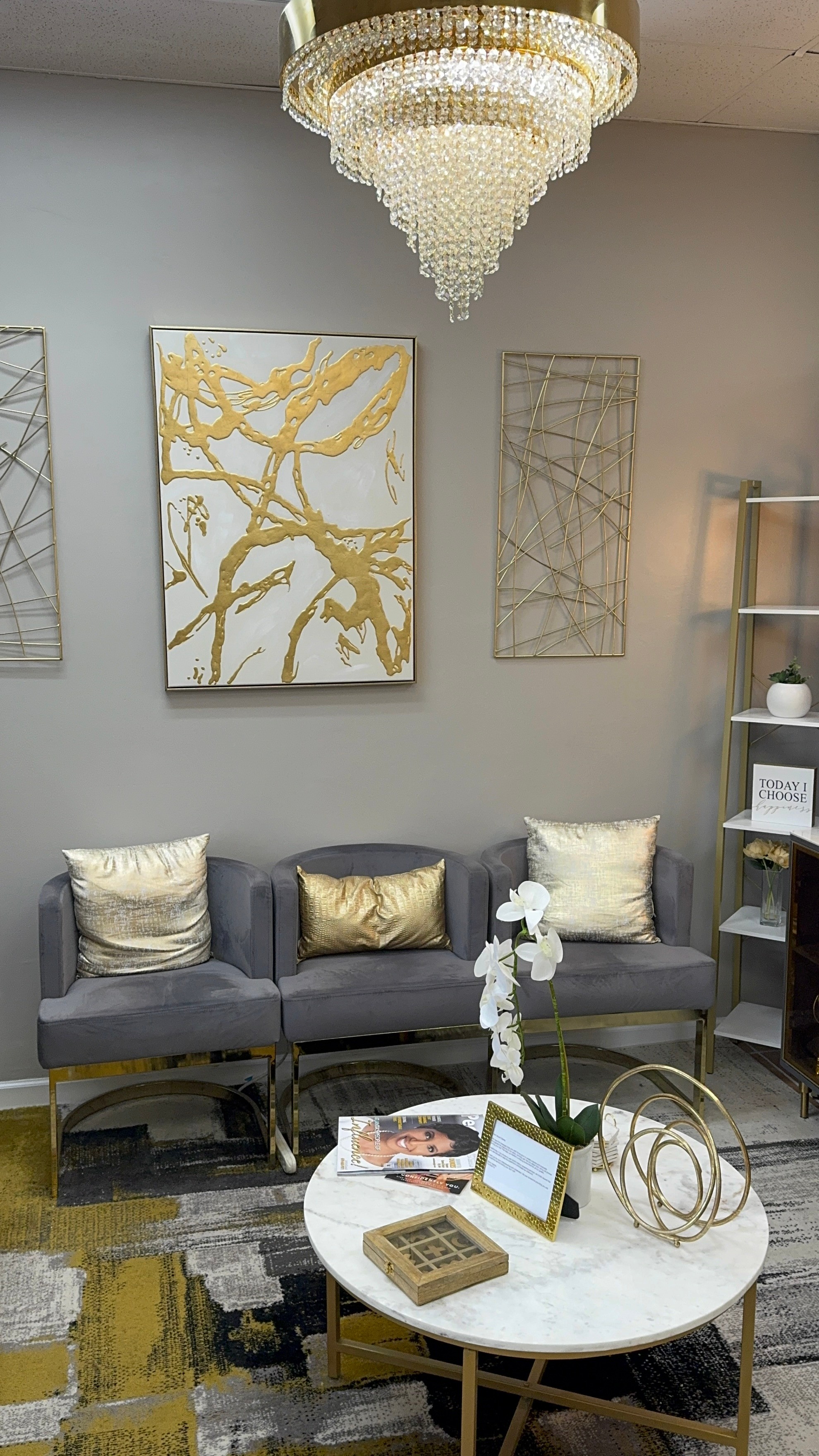 Living room with gray sofa, gold and white pillows, white round table, chandelier, and wall art, with a bookshelf nearby.