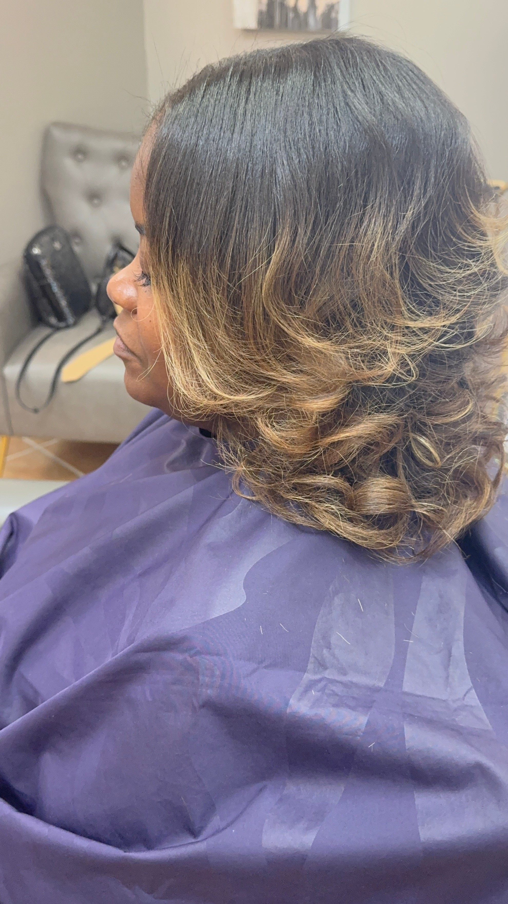 Side view of a woman with shoulder-length wavy hair, wearing a purple cape, sitting in a salon chair.