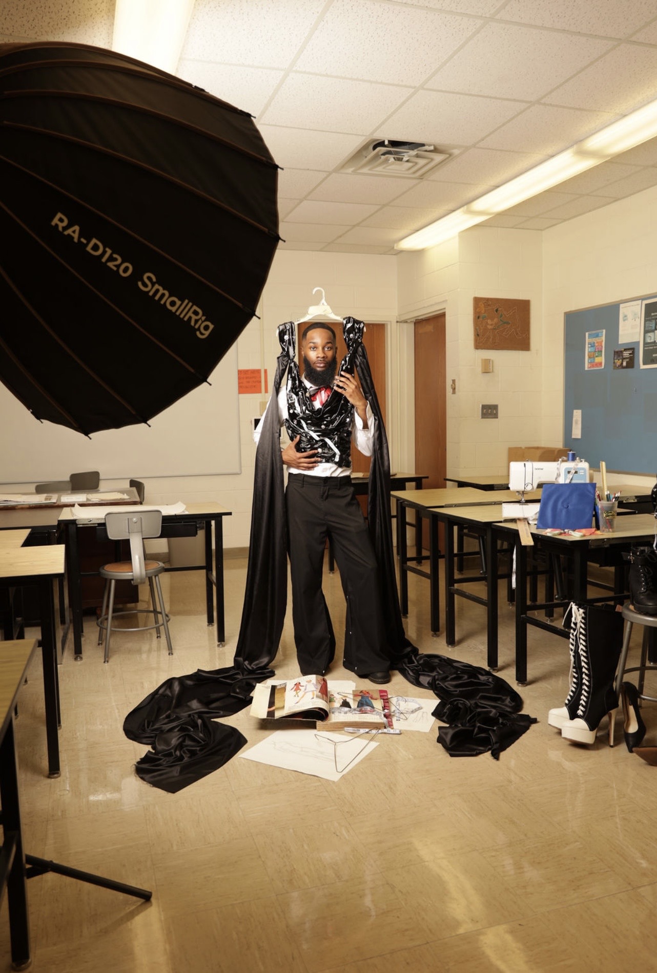 Person taking a mirror selfie in a classroom, surrounded by tables, chairs, and a large softbox light, with clothes on the floor.