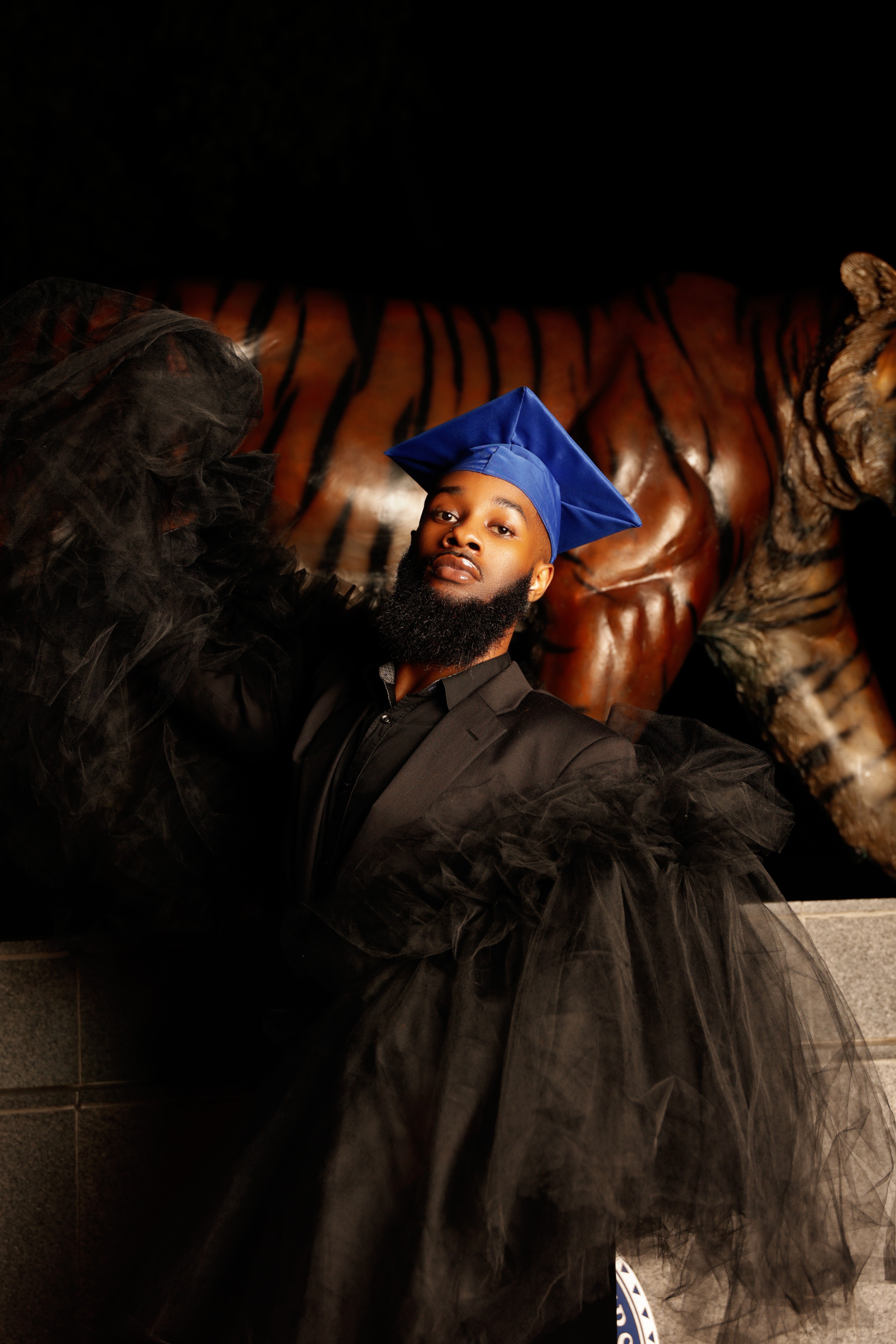 Person in graduation cap and gown sitting on a tiger statue, with a dark background.