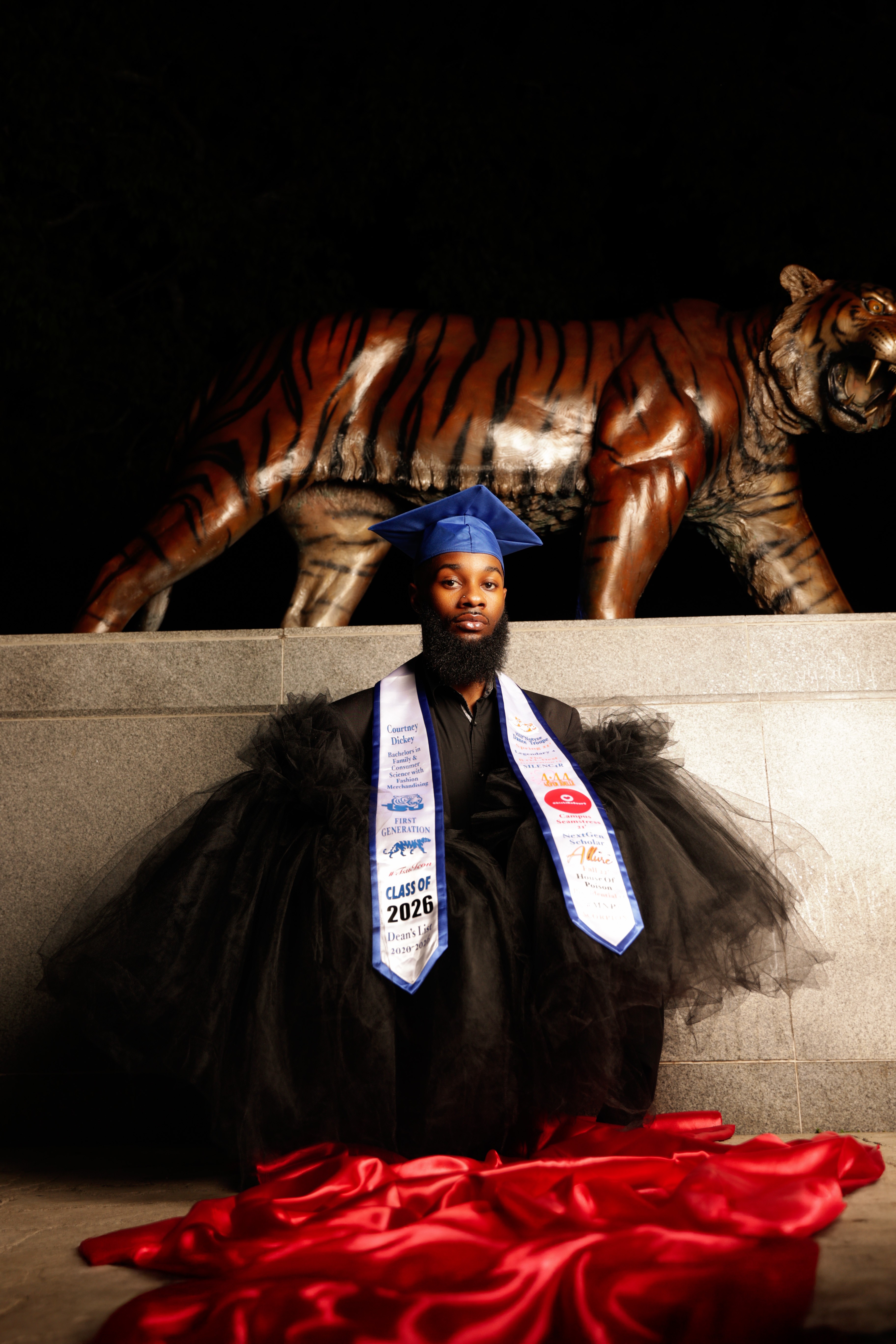Person in a black dress with a blue cap and graduation sash, standing in front of a tiger sculpture, with red fabric on the ground.