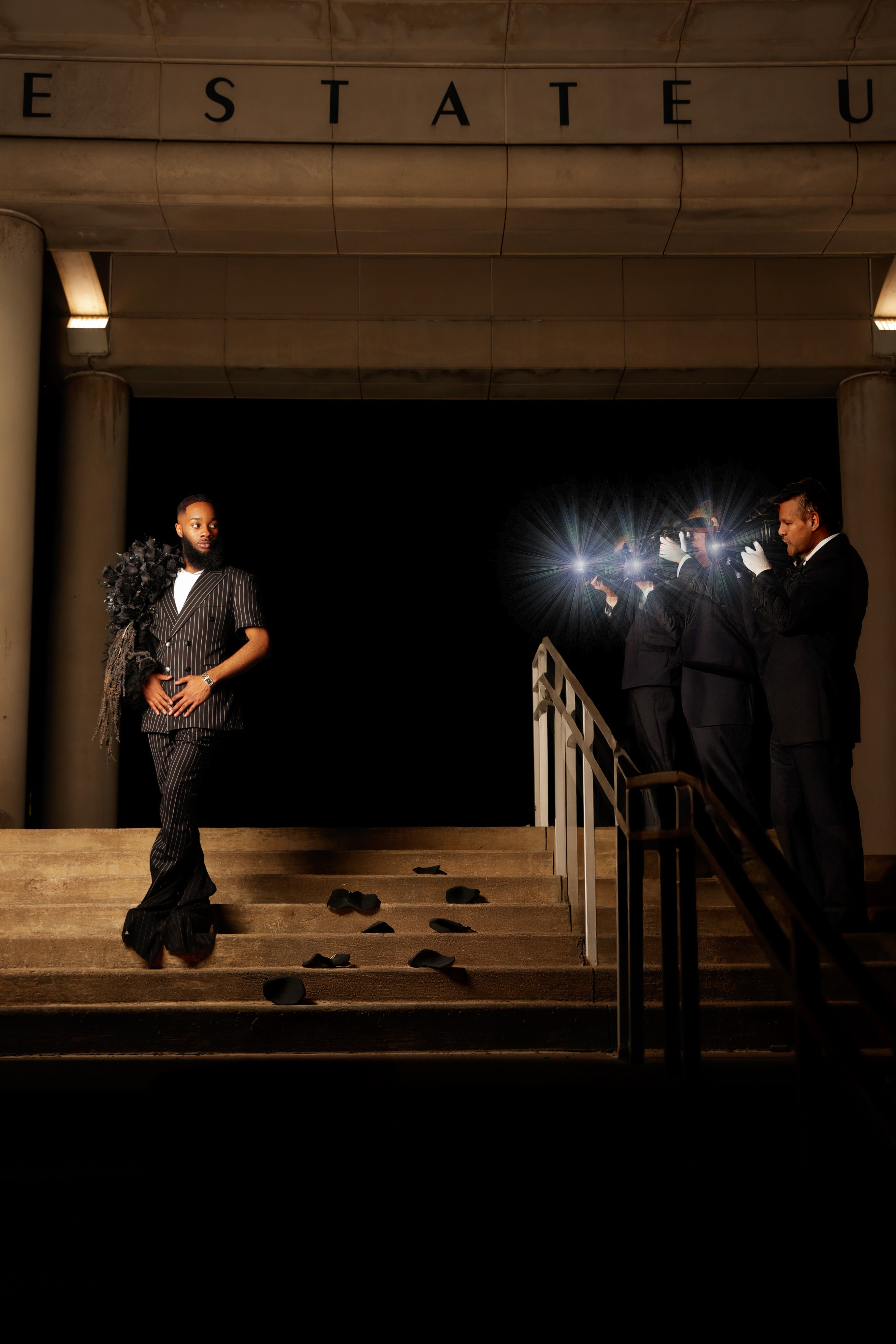 Four people in formal attire on stage, two facing each other, one standing alone, with a dark background and bright light behind the group.