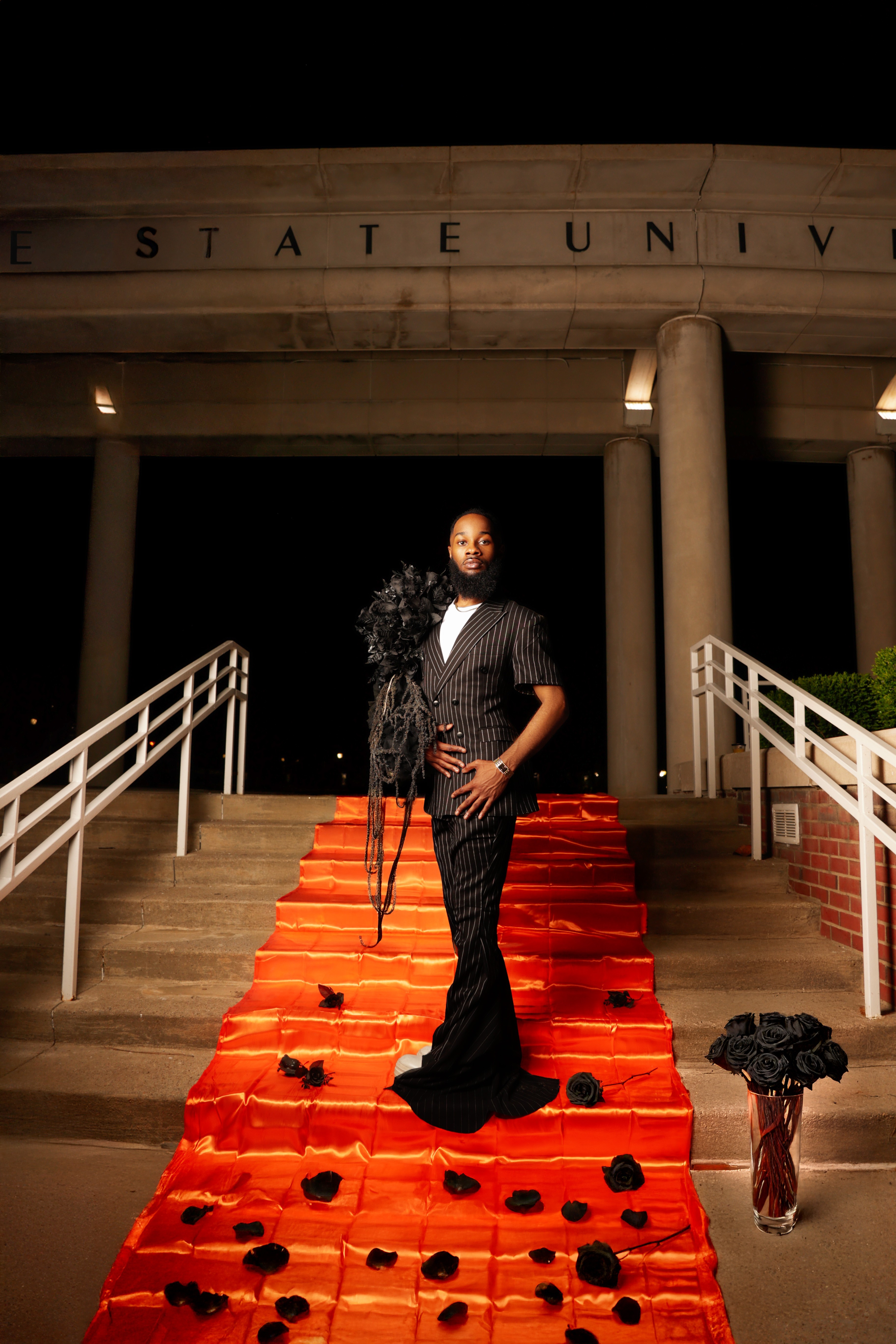 Person standing on a red carpet with black flowers, in front of a building with columns and partially visible text, at night.